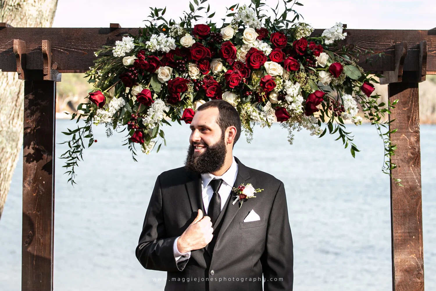 Man in a black suit with a beard stands under a wooden pergola adorned with red and white roses near a lake.