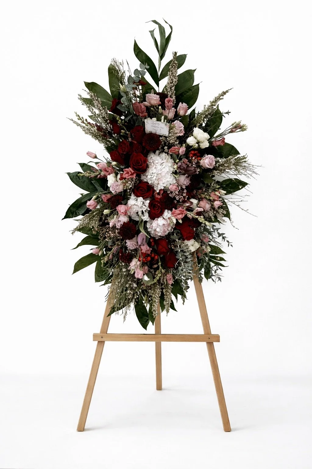 Colorful floral arrangement with roses, hydrangeas, and greenery displayed on a wooden easel against a white background.