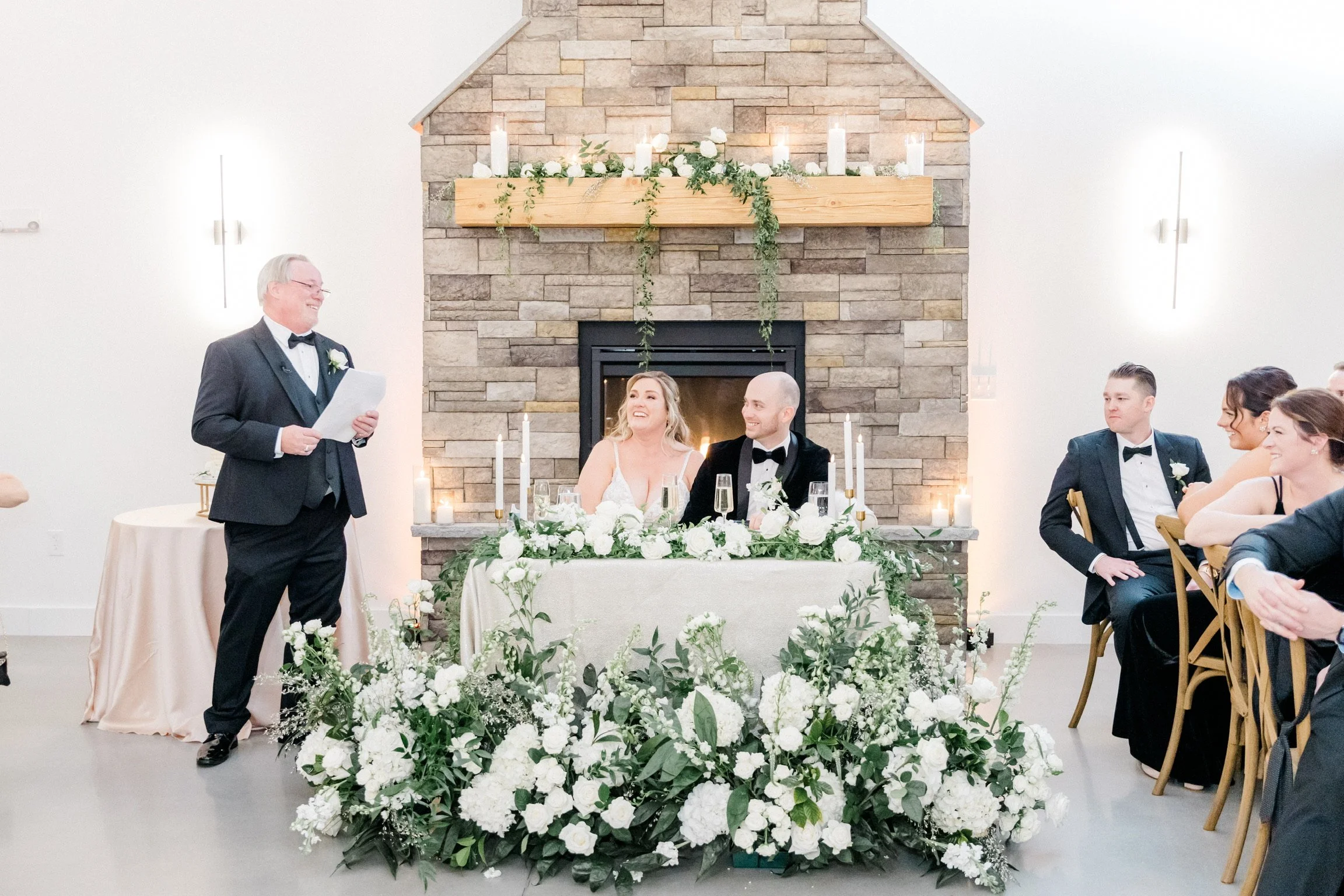 Bride and groom at wedding reception table with guests and man giving speech, surrounded by white floral arrangements and candles.