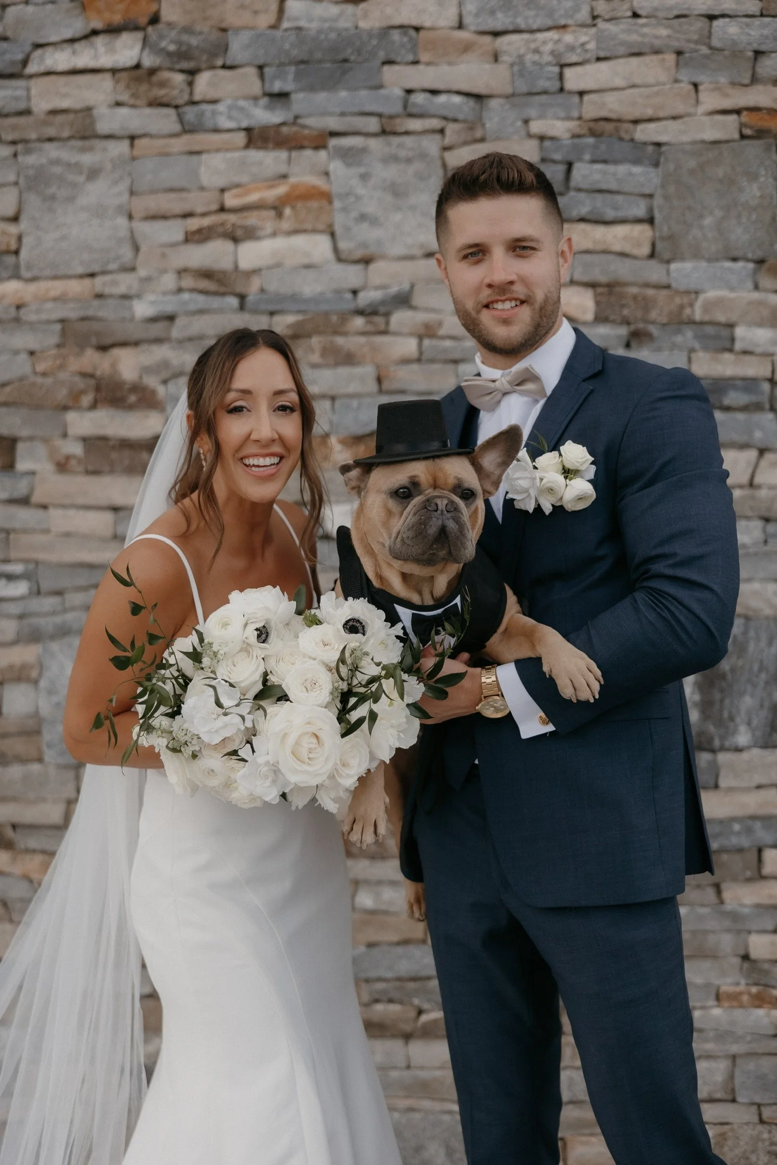 Bride and groom posing with a French Bulldog wearing a top hat in front of a stone wall. The bride is holding a bouquet of white flowers.