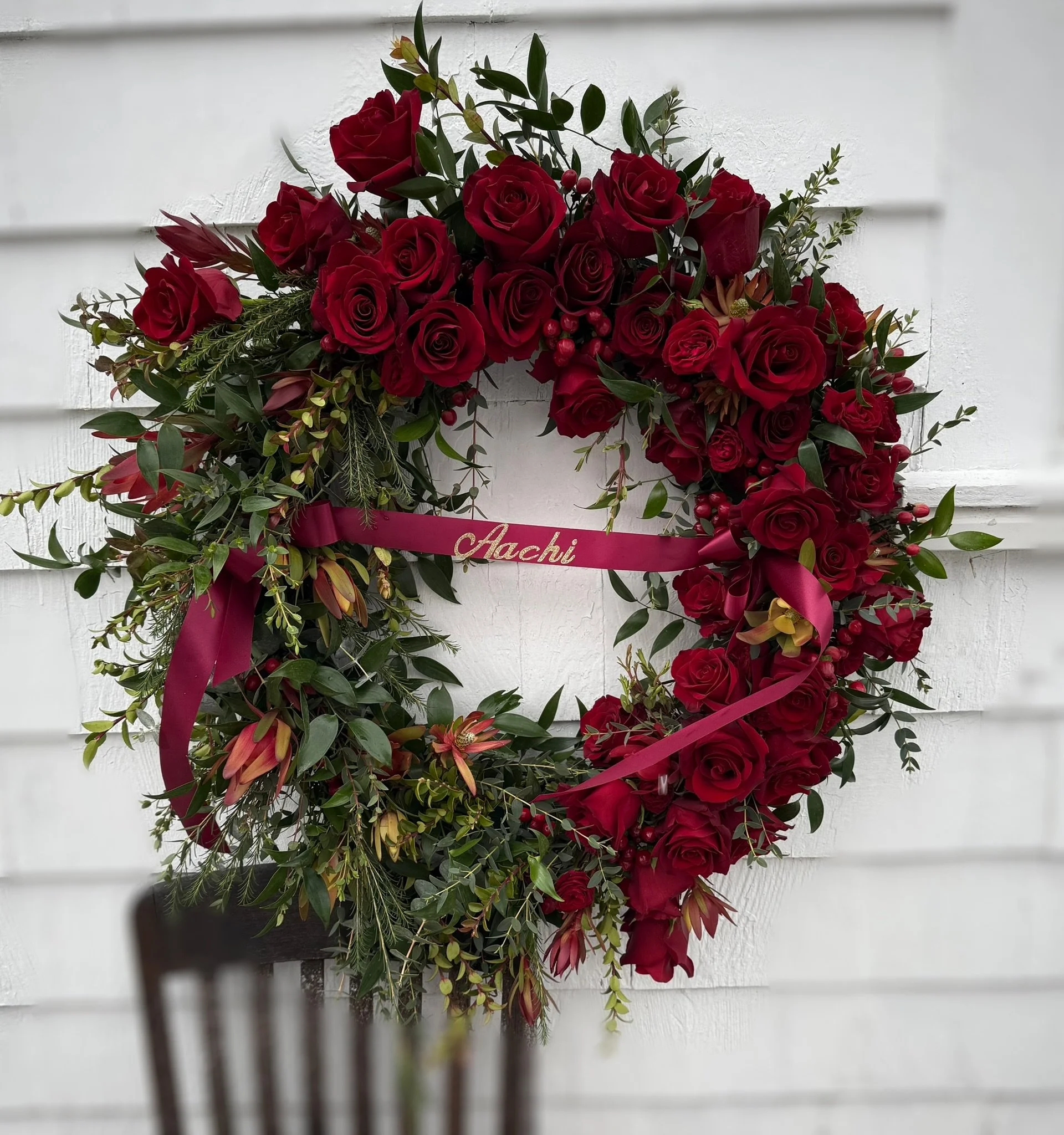 Wreath of red roses and greenery with a maroon ribbon inscribed 'Aachi' hanging on a white wall.