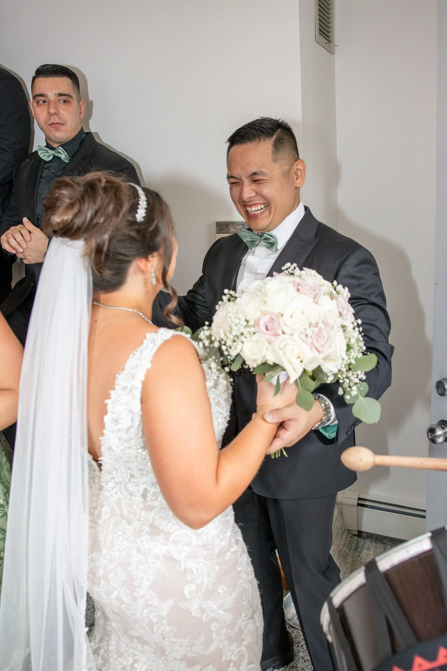 A bride in a white lace gown holding a bouquet of white and pink roses exchanging smiles with a groom in a dark suit and bow tie, both appearing joyful, with a groomsman in the background.