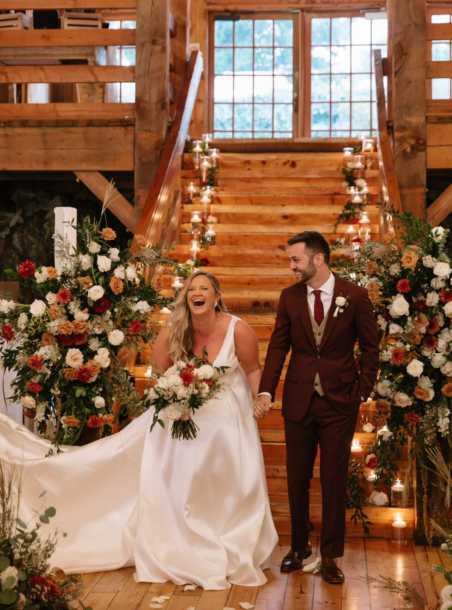 Smiling bride and groom at rustic indoor wedding with floral arrangements and candles on wooden staircase.