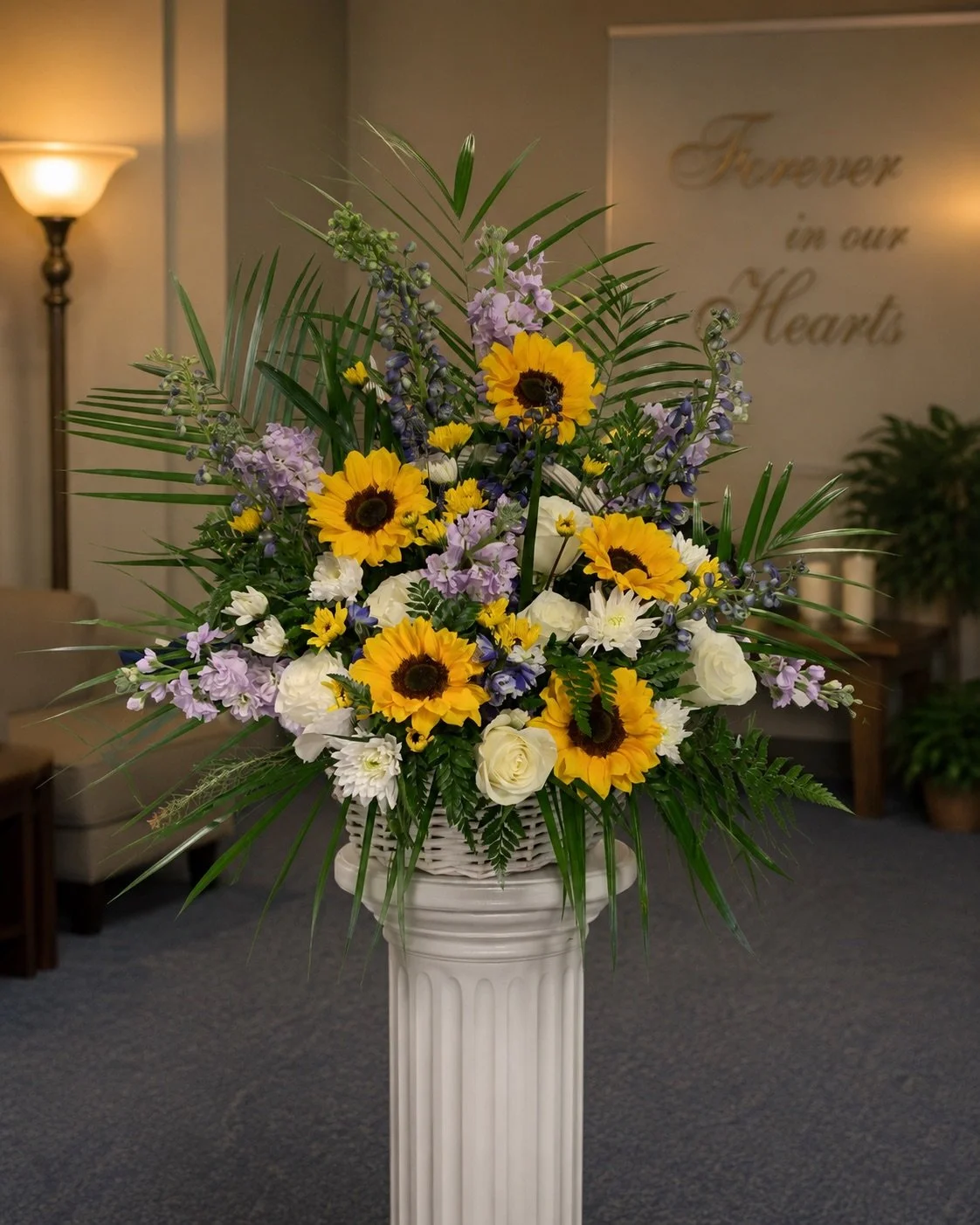 A floral arrangement with sunflowers, white roses, and purple and white flowers, in a white vase on a pedestal, in a room with soft lighting and a sign that reads 'Forever in our Hearts' in the background.
