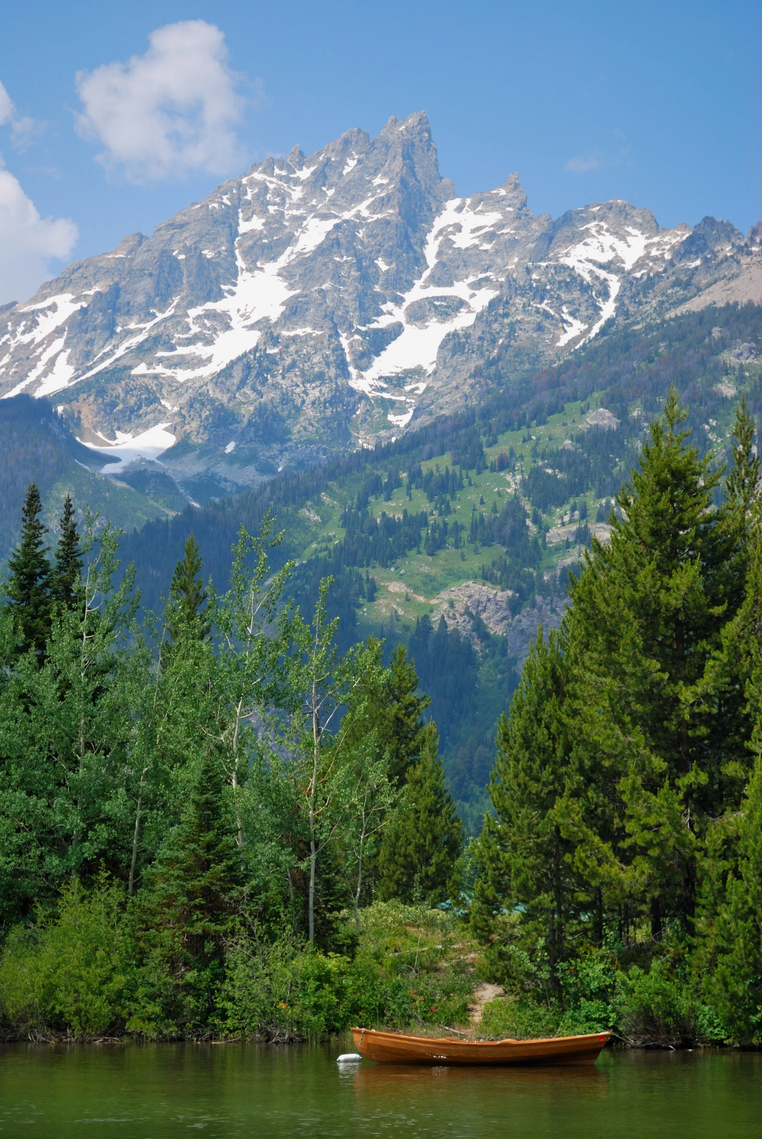 A mountain with snow patches in a lush green forest, a calm lake in the foreground, and a small boat floating on the water.
