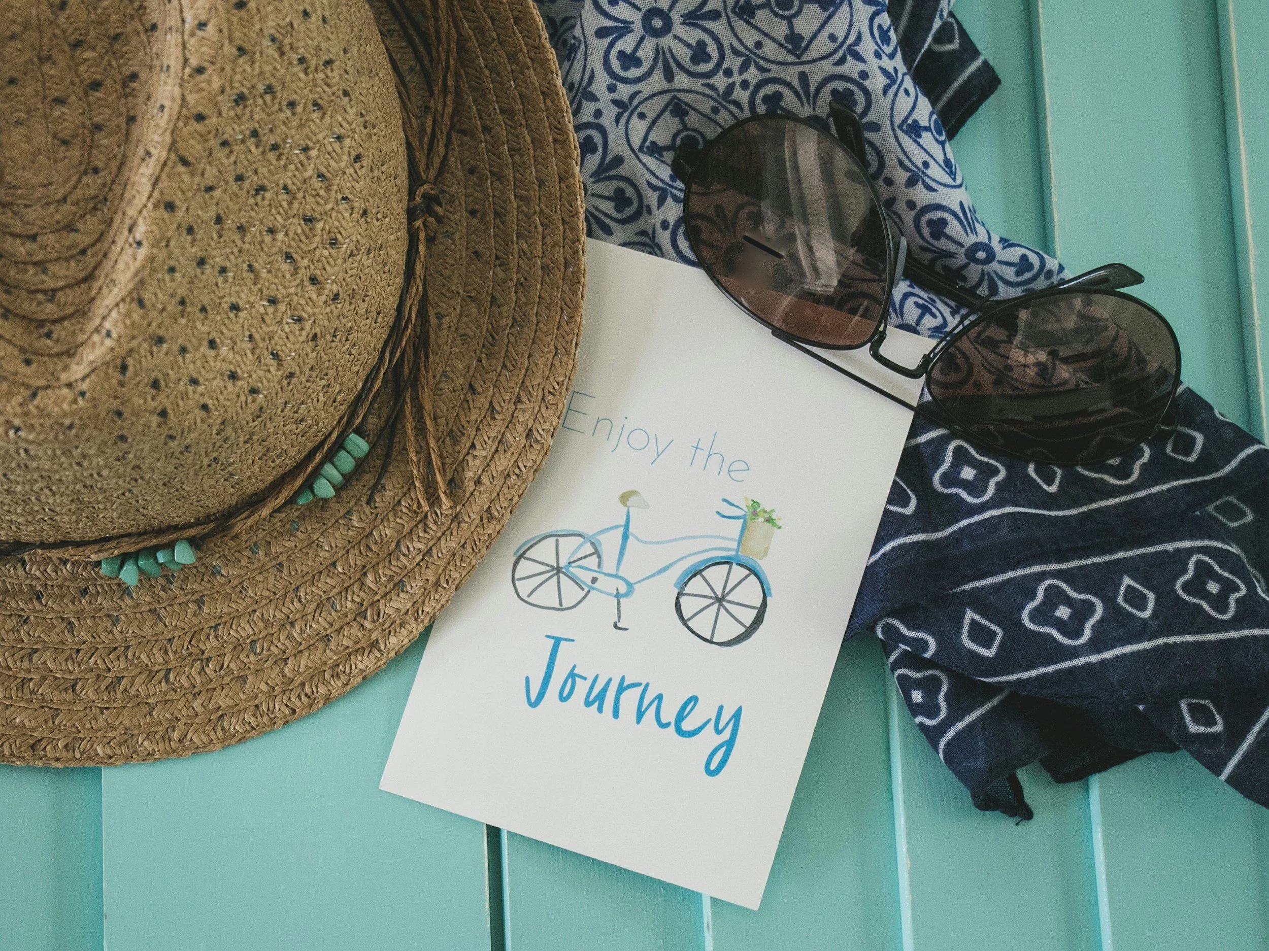 A woven straw hat with turquoise beads around the brim, sunglasses with dark lenses, a piece of paper with a bicycle illustration and the words "Enjoy the journey," a blue and white patterned cloth, and a dark bandana on a turquoise table.