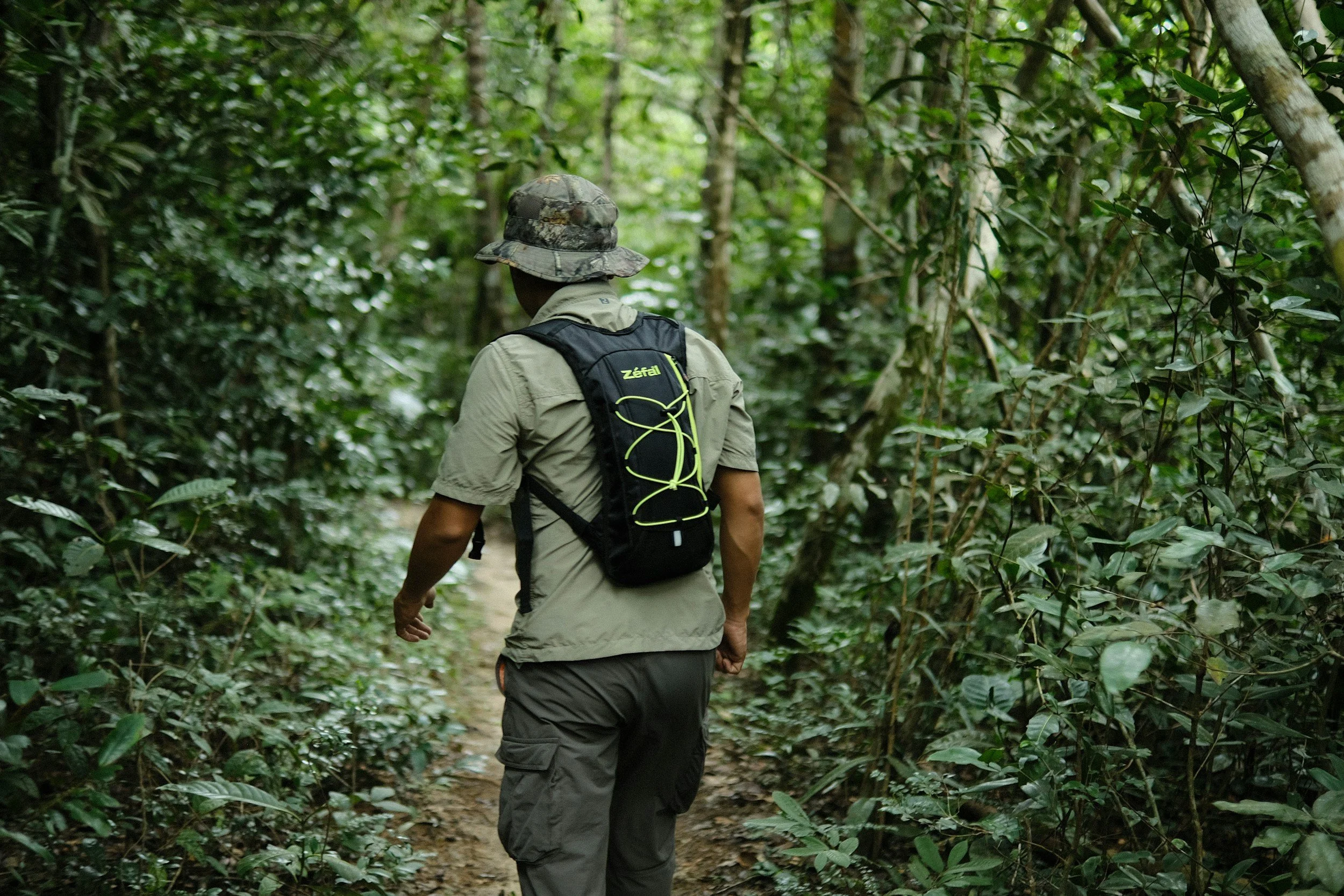 A person wearing a camouflage hat, khaki shirt, and cargo pants walking through a dense green forest with a backpack.