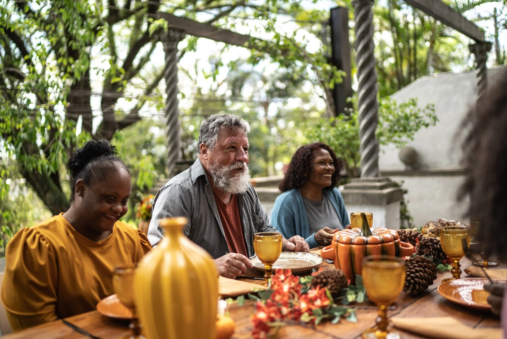 Group of diverse people enjoying a meal outdoors at a table decorated with fall-themed decorations, surrounded by trees.