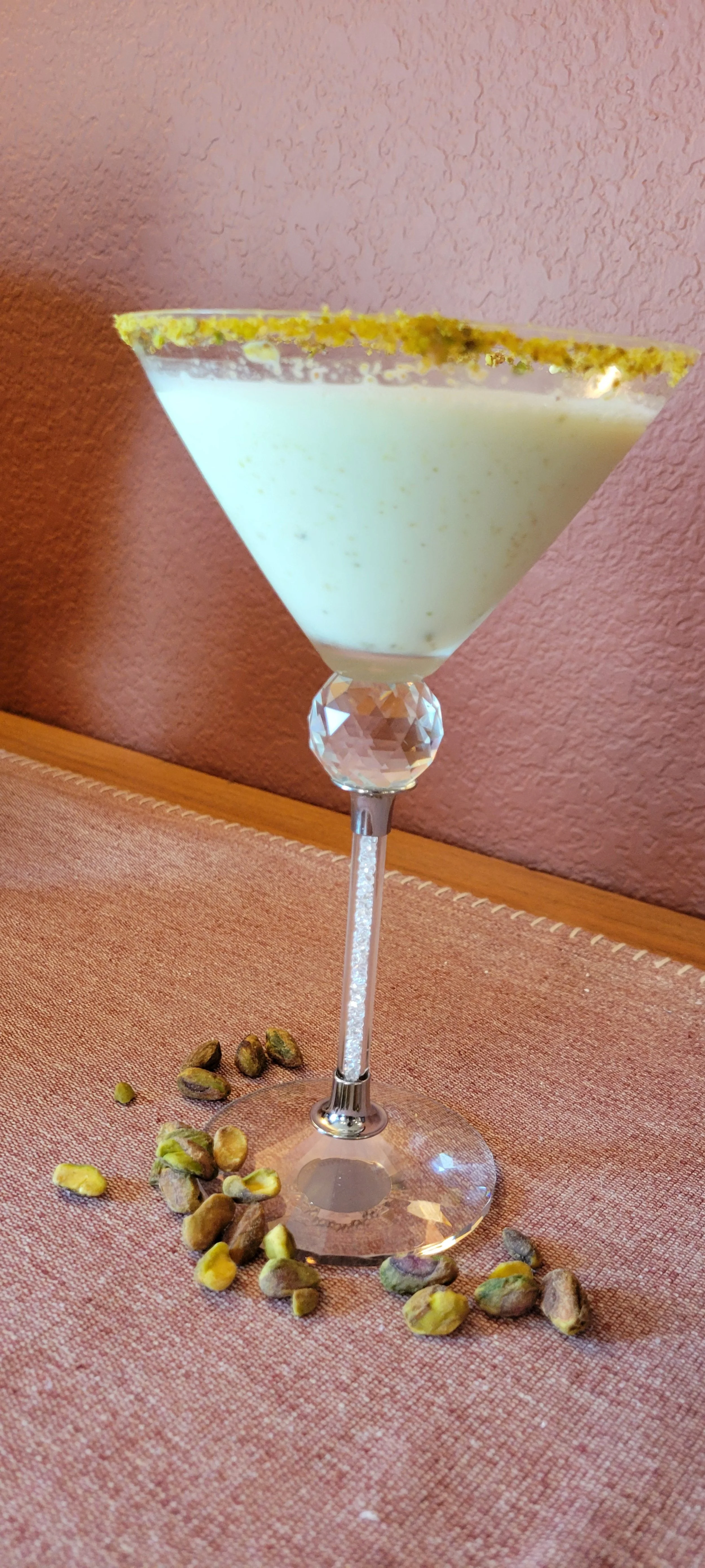 A green pistachio-flavored dessert in a decorative martini glass, garnished with crushed pistachios on the rim, with whole pistachios scattered on a pink textured tablecloth.