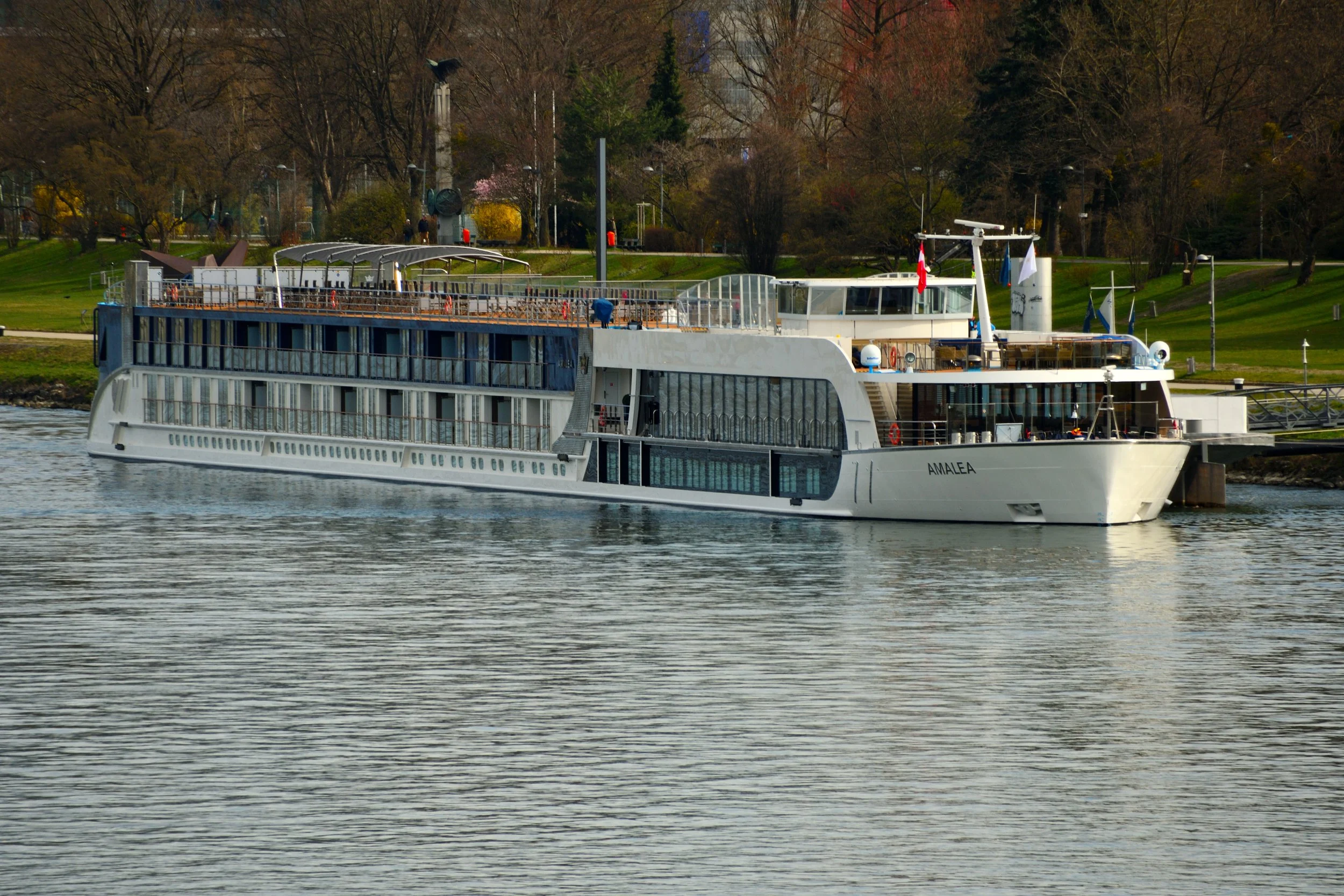 A white multi-deck boat named Amalea sailing on a river with a green park and leafless trees in the background.
