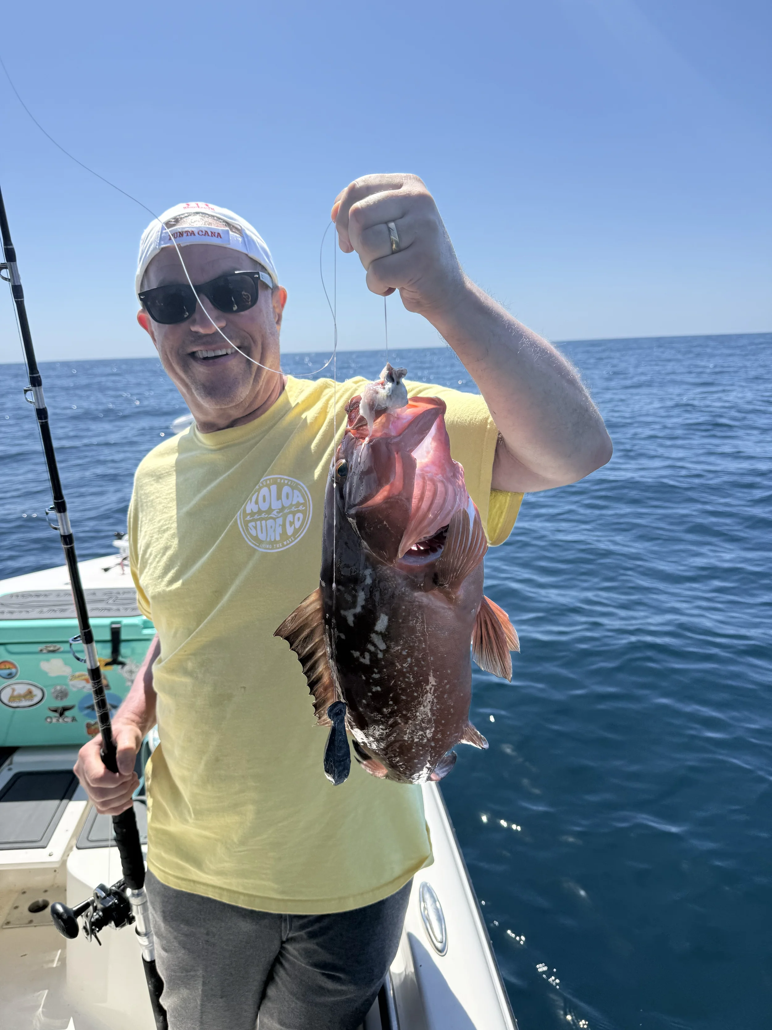 A man in sunglasses, a white cap, and a yellow T-shirt holding a fishing rod and smiling while holding a large fish on a boat in the ocean during a sunny day.