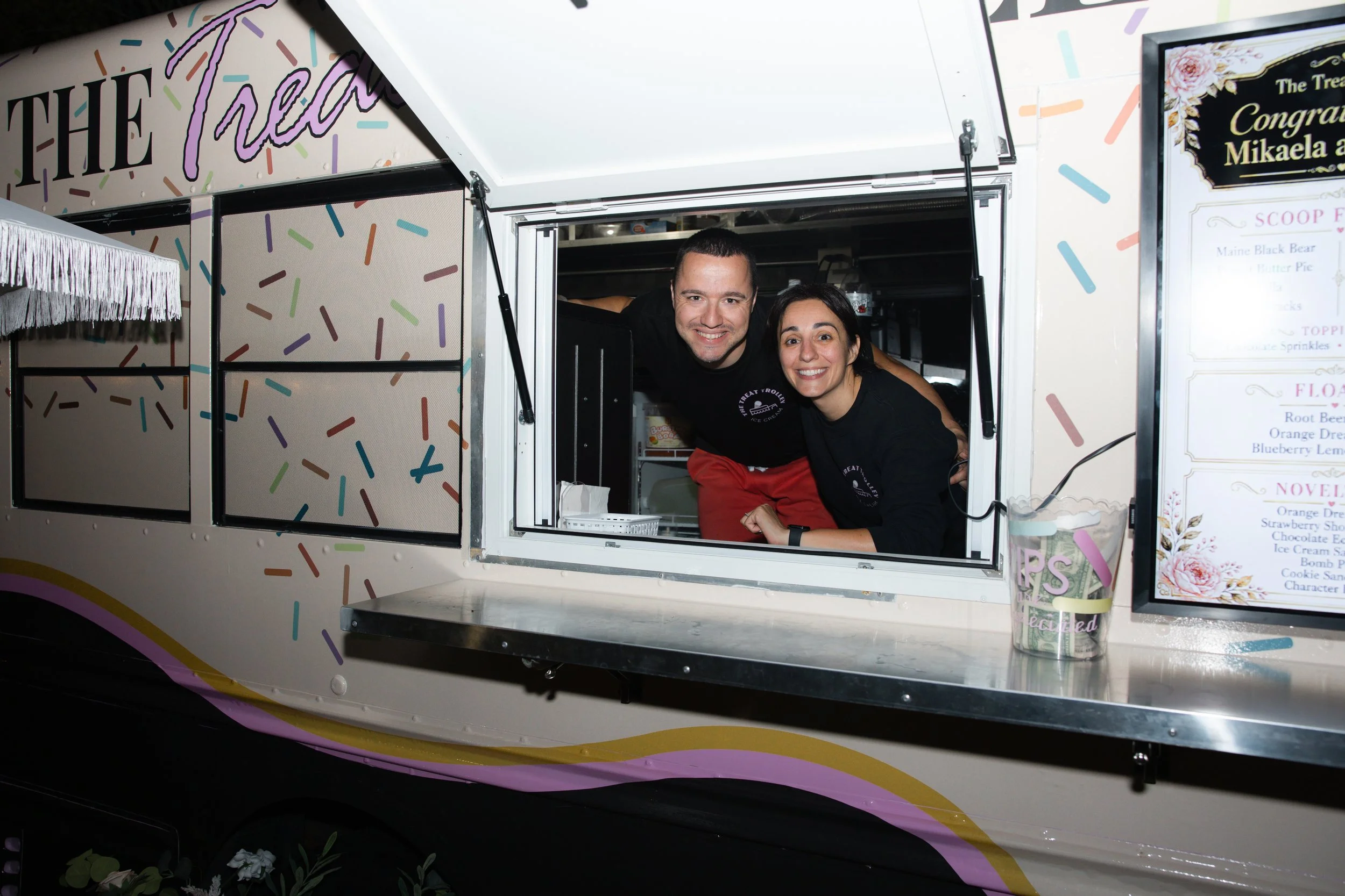 Two smiling people leaning out of a colorful ice cream truck window, with a menu and decorative confetti design on the truck.