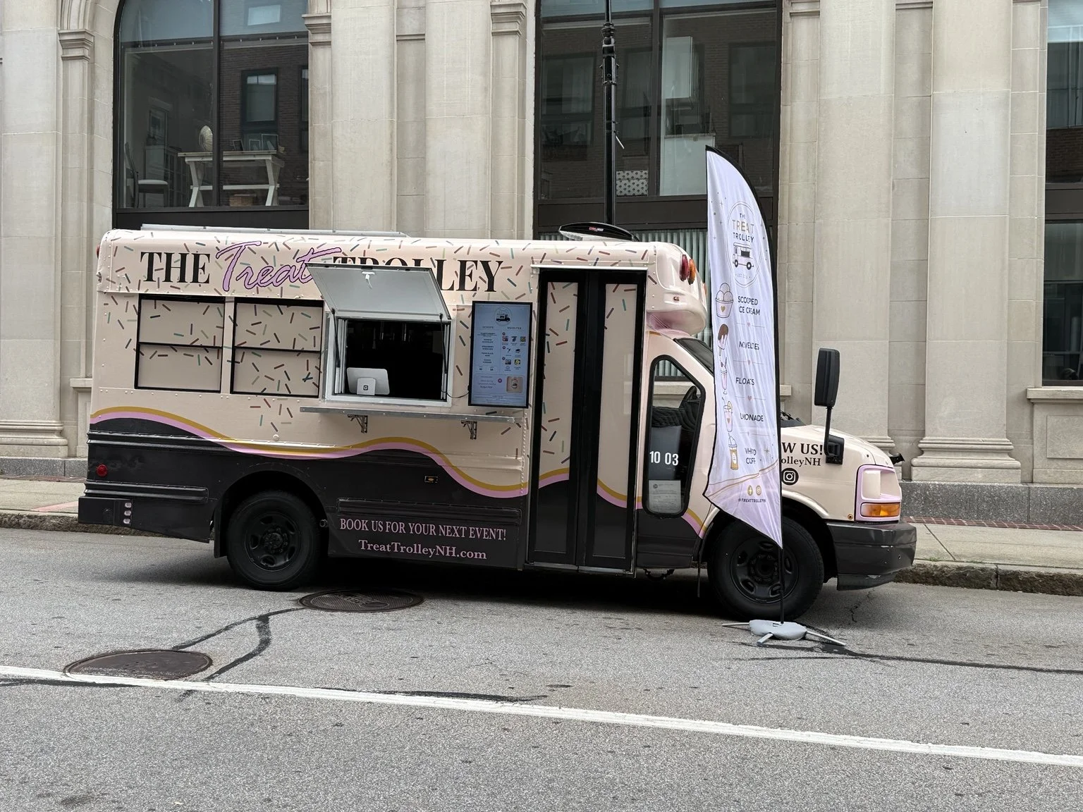 A pink and black Treat Trolley food truck parked on the street in front of a large stone building with tall windows. The truck has colorful sprinkles graphics, an order window, and a tall banner flag displaying menu items like scooped ice cream, smoo