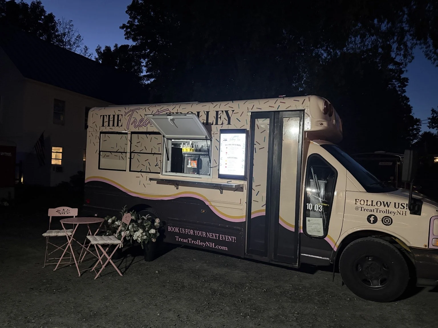 A food truck decorated in pastel pink and yellow wavy stripes, with seating outside including two pink chairs and a small table. The truck has the words 'The Treat Trolley' on the side, a menu display, and social media information. It is parked outsi