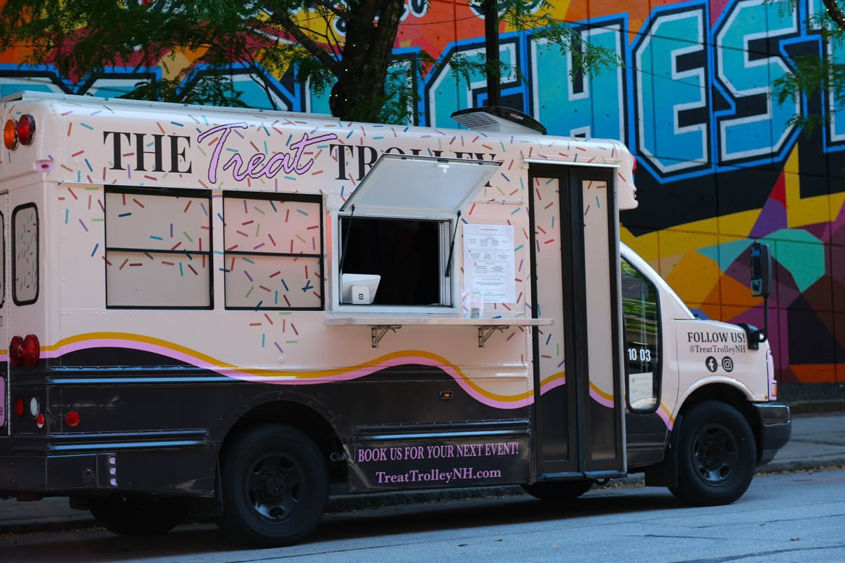 A pink ice cream truck named 'The Treat Trolley' is parked on a city street with a colorful graffiti mural in the background. The truck has a serving window open, and the text on the side encourages booking for events and following on social media.