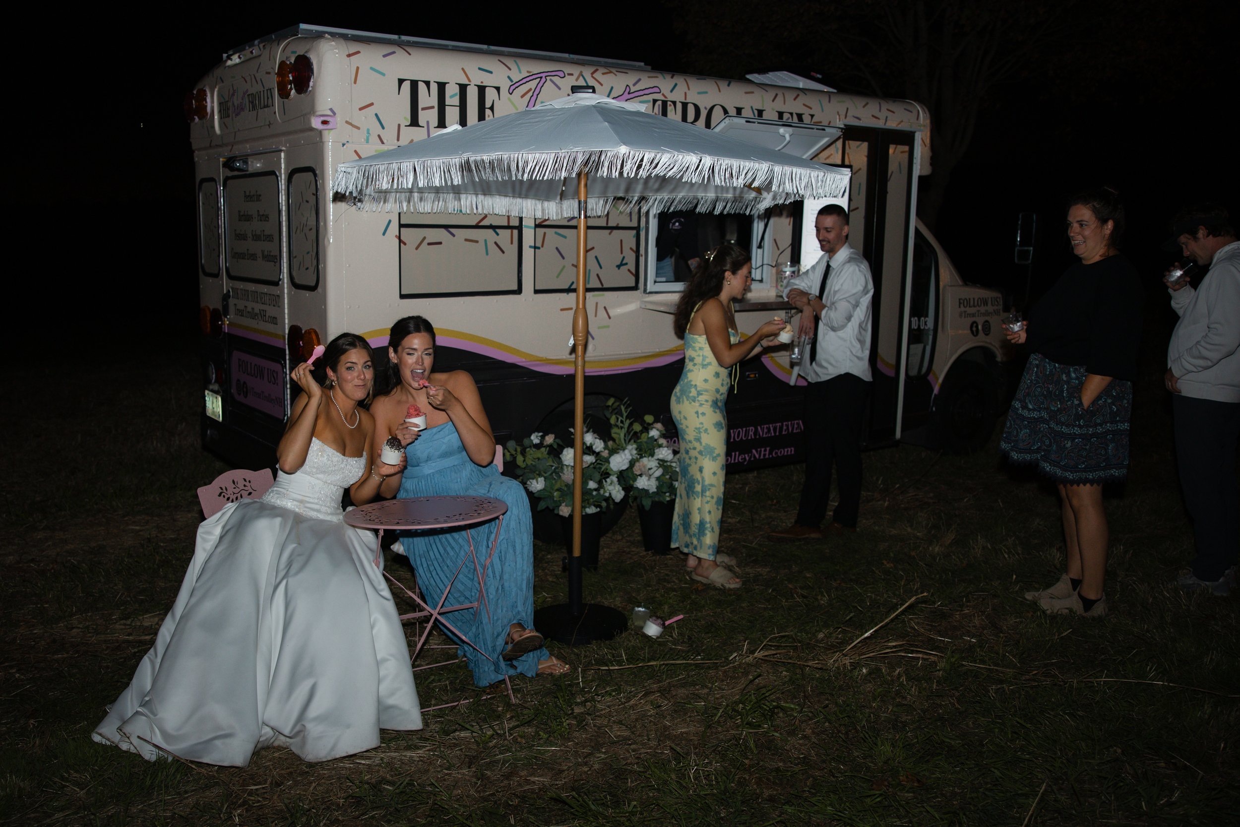 Group of people celebrating outdoors at night near an ice cream food truck, with two women in wedding dresses, one enjoying ice cream and the other sitting on a pink chair, and others chatting nearby.