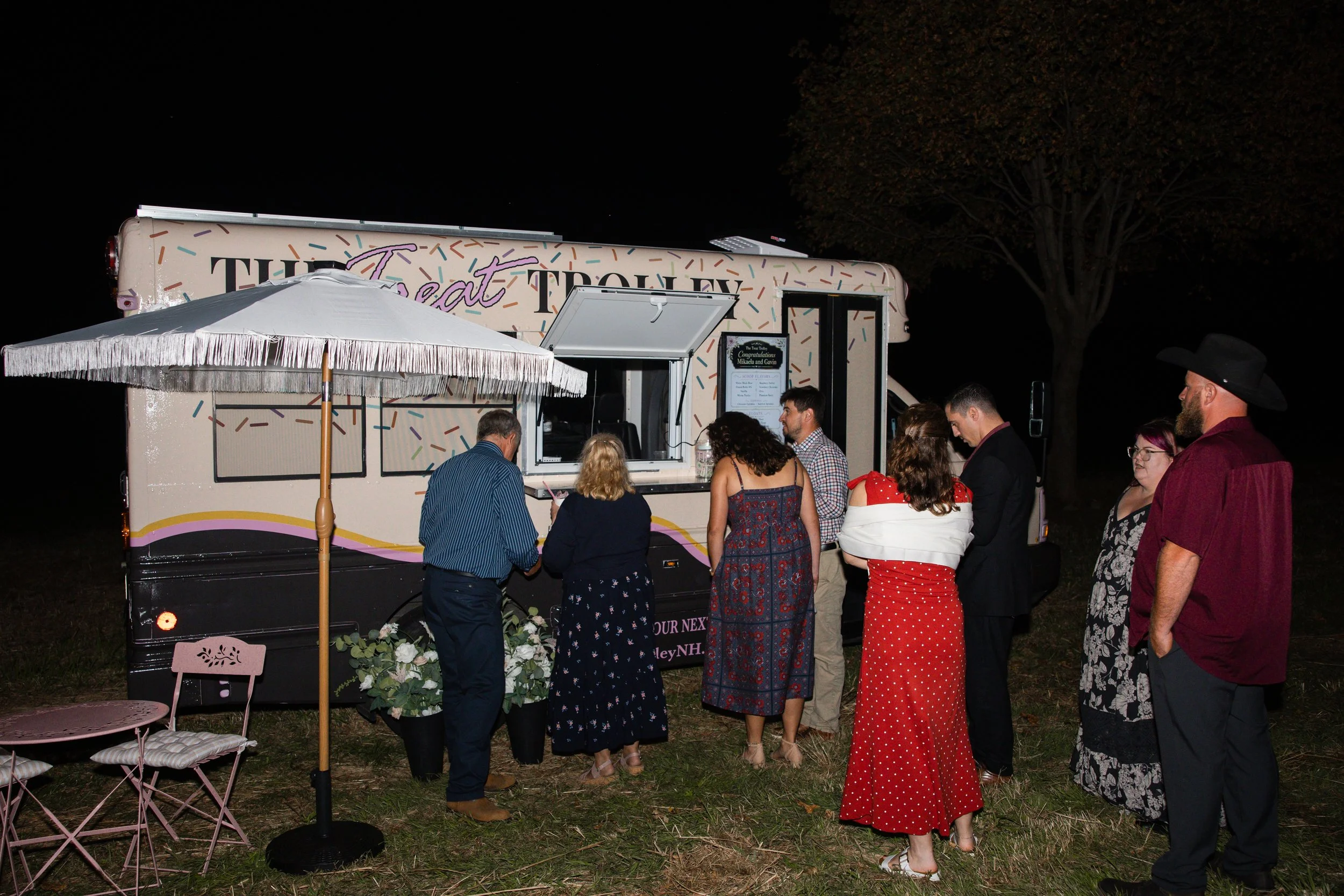 People gathered outside at night near a decorated mobile food truck, with an umbrella, pink chairs, and a tree in the background.