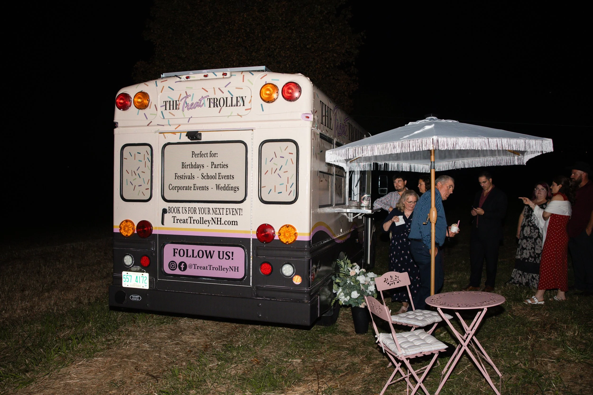 Night scene with a decorated ice cream truck labeled "The Treat Trolley" and people gathered around, under a white umbrella with pink chairs nearby.