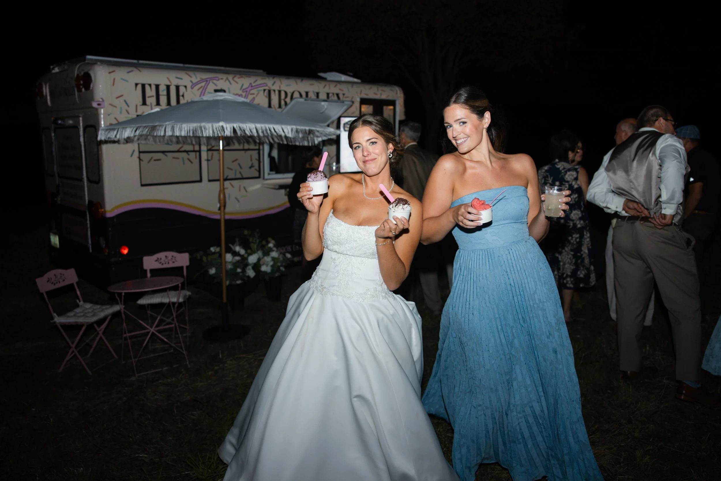 Two women in formal dresses smiling and holding ice cream and a drink at night, with a food truck and other people in the background.