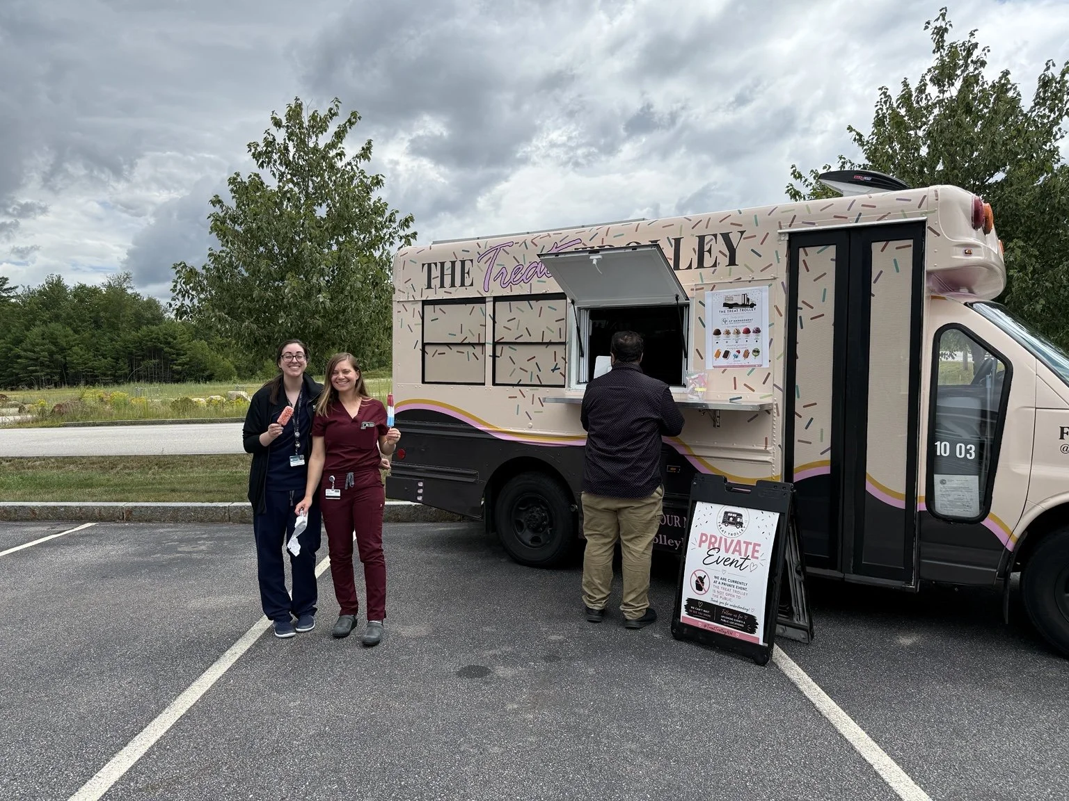 Two women standing and smiling in front of a mobile ice cream truck, with a man ordering at the window, in an outdoor parking lot with trees and cloudy sky in the background. Corporate Outing