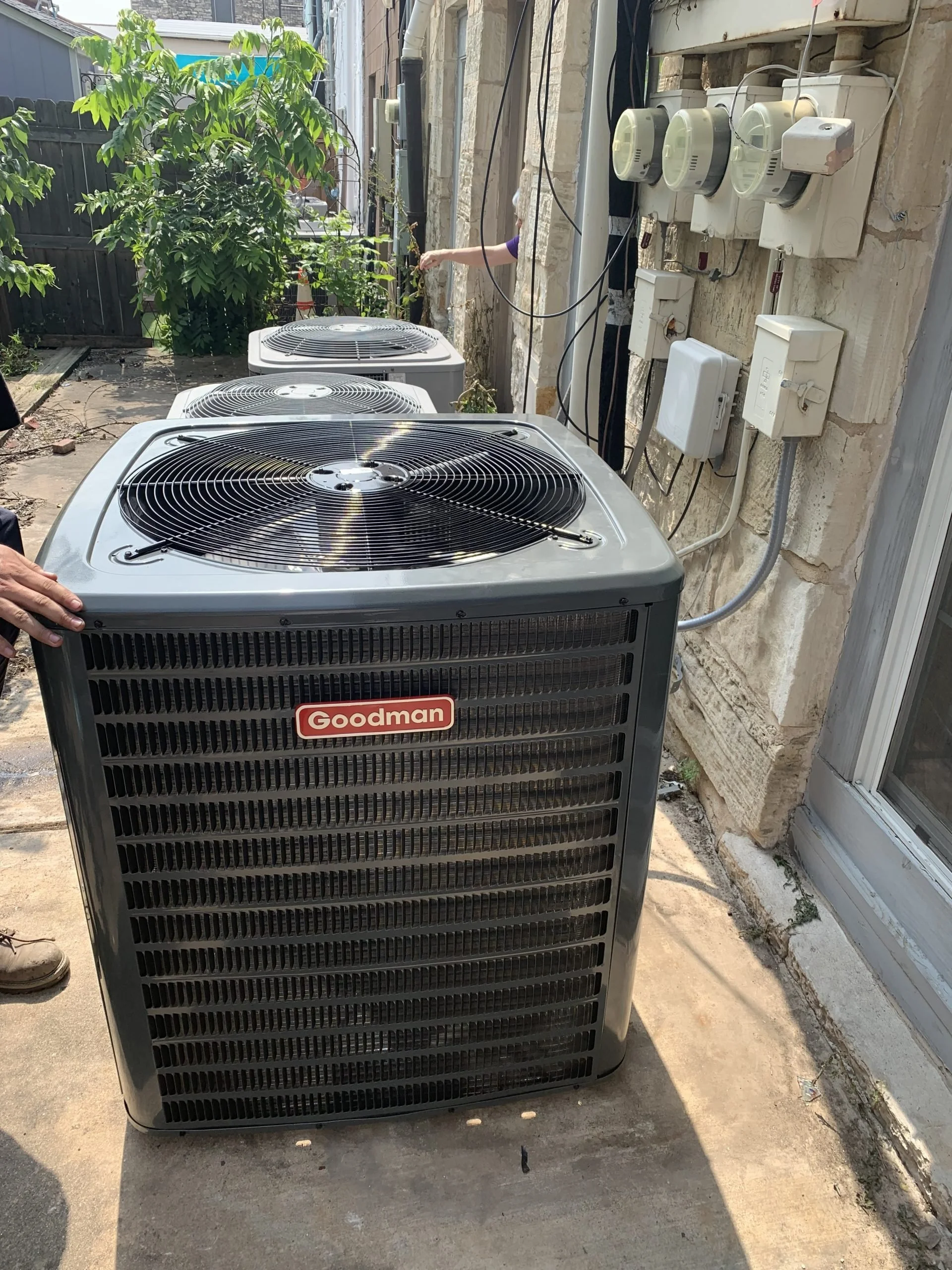 Three Goodman outdoor air conditioning units placed on a concrete surface outside a building, with electrical and communication boxes on the wall nearby.