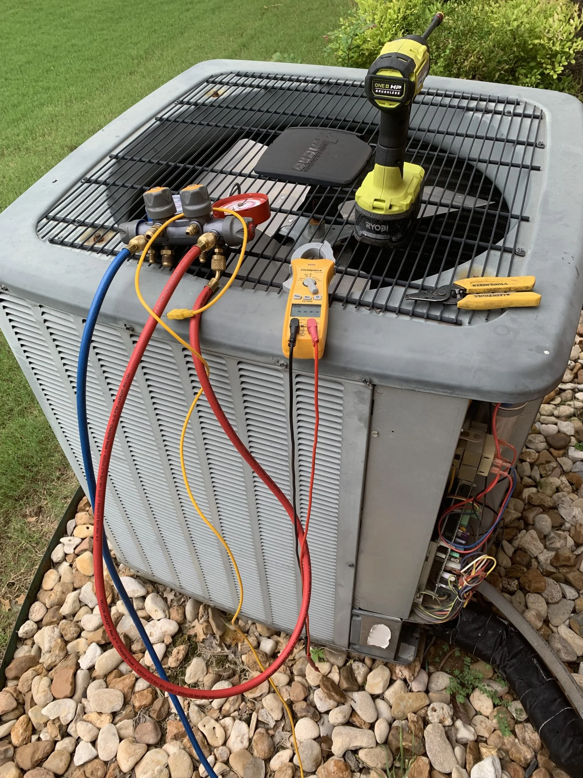 An outdoor air conditioning unit being serviced with tools and electronic testing devices on top, including refrigerant hoses and a digital multimeter.
