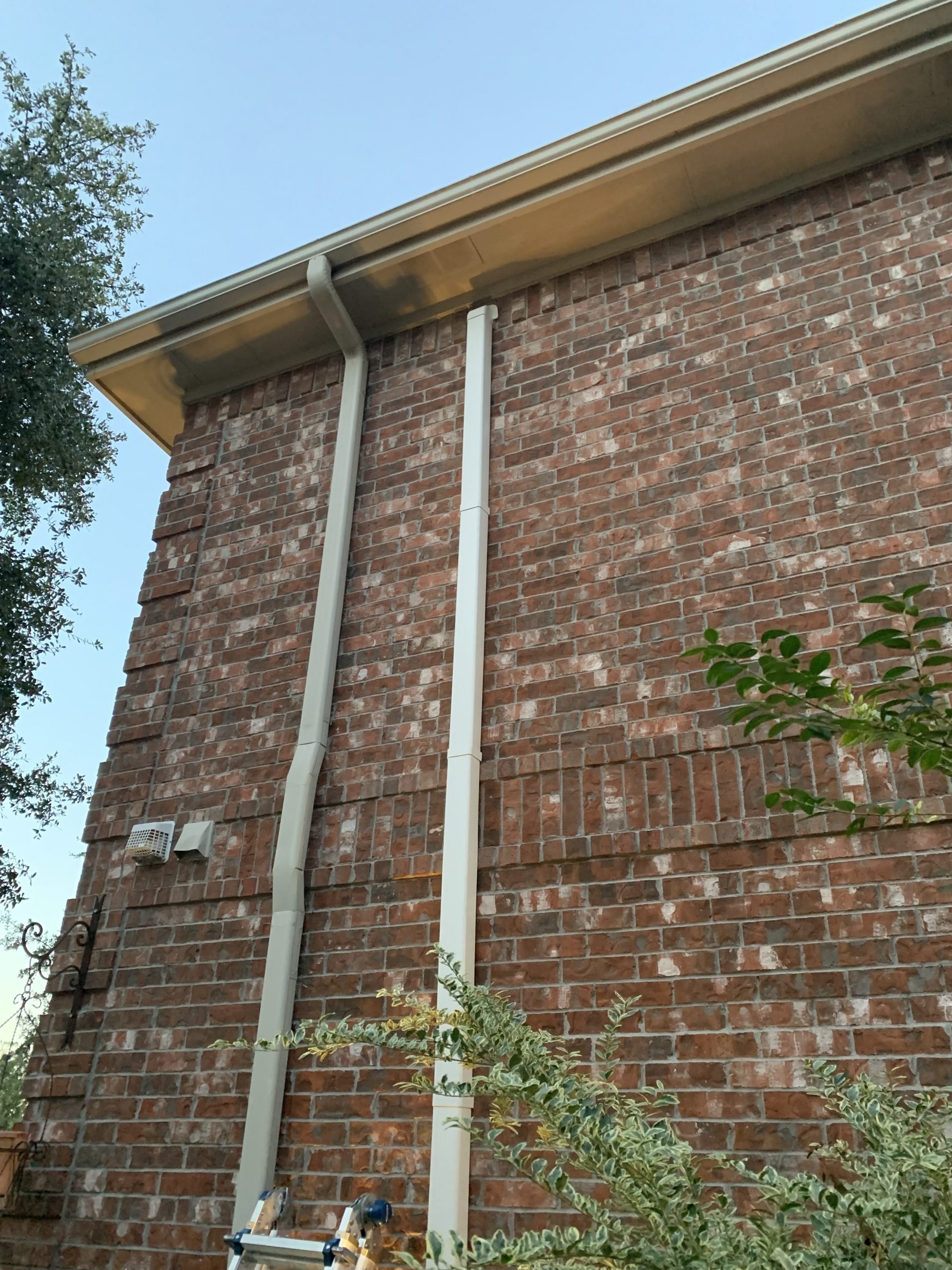 Close-up of a brick house exterior showing white gutter pipes running down the wall, with some greenery at the bottom and a small vent and security light on the wall.