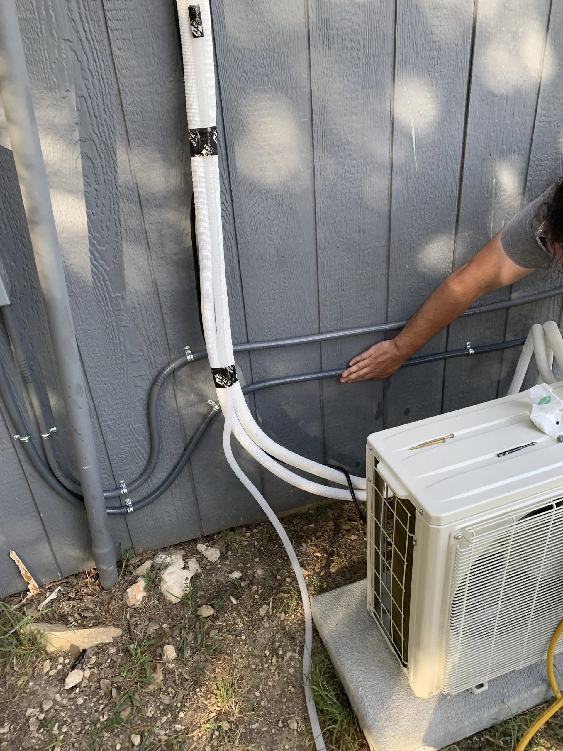An outdoor HVAC unit connected to insulated refrigerant lines and electrical conduit on the side of a building, with a person installing or servicing the system.