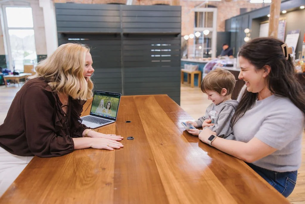 Two women and a young boy sitting at a wooden table in a cafe, smiling and laughing. The woman on the left has blonde hair, and the woman on the right has dark hair. The boy is holding a smartphone. The woman on the left has a laptop open in front of her.