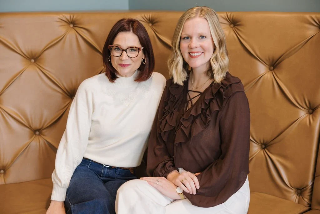 Two women sitting in front of a tufted leather headboard, smiling at the camera.