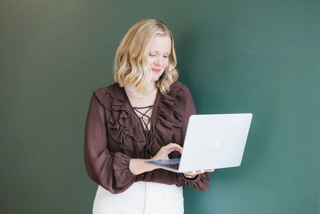 A woman with blonde hair wearing a dark ruffled blouse and white pants, standing against a green wall, looking at a laptop.