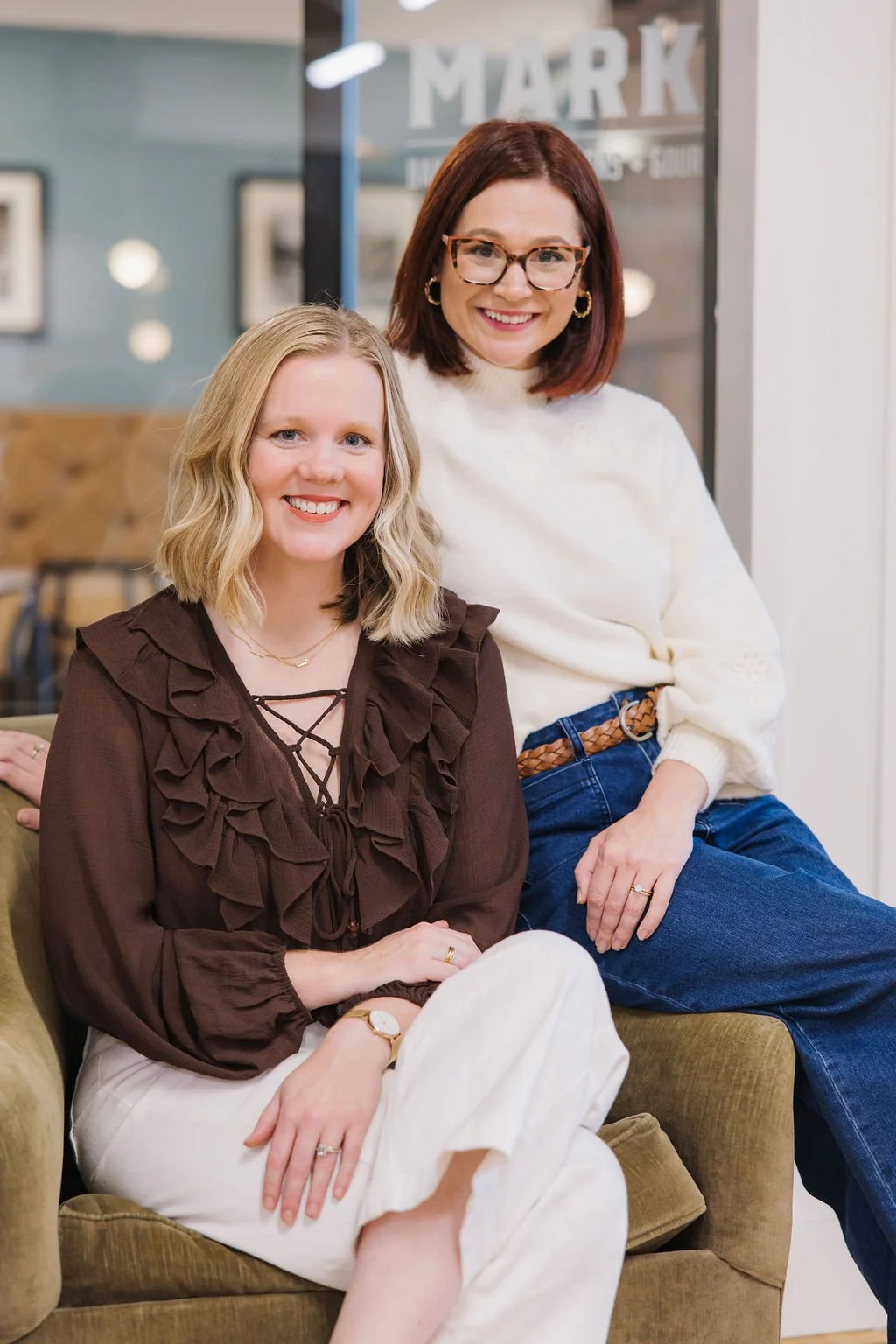 Two women sitting on a green couch inside a cozy, modern café with artwork on the walls. The woman on the left has blonde hair, a brown top with ruffles, cream pants, and is smiling at the camera. The woman on the right has shoulder-length dark red hair, glasses, a white sweater with embroidered details, jeans, and is also smiling.