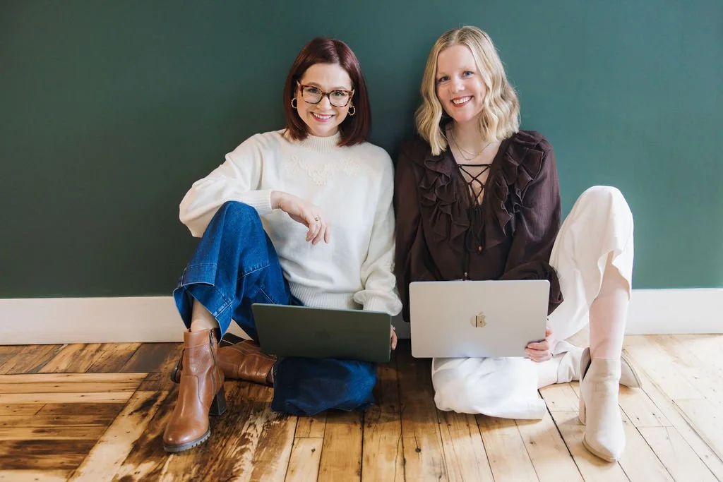 Two young women sitting on a wooden floor against a green wall, smiling at the camera, each using a laptop.