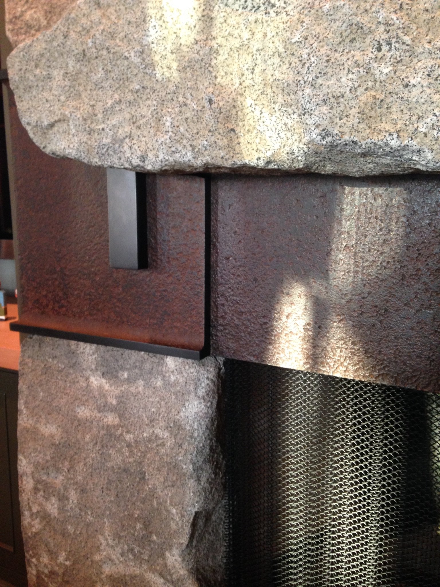Close-up of a fireplace with a granite stone mantel, rust-colored metal panel, and textured stone and metal surfaces.