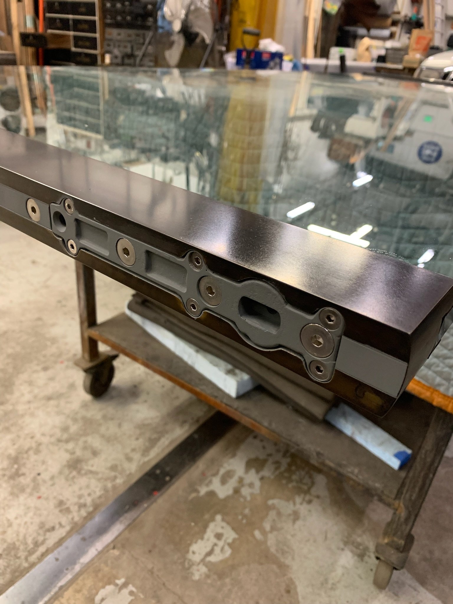 Close-up of a metal frame or assembly with multiple screw holes, placed on a wooden workbench in a workshop setting.