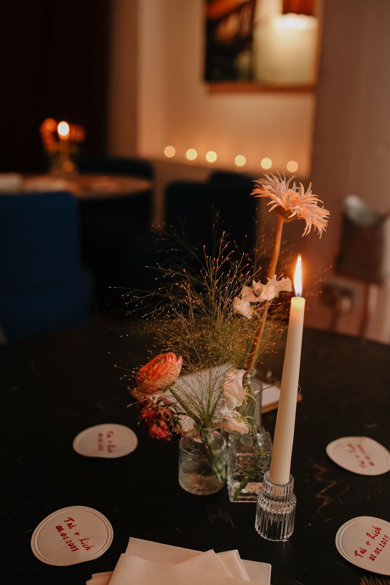 A table centerpiece with flowers and a lit candle in a glass holder at a dimly lit celebration or dinner event.
