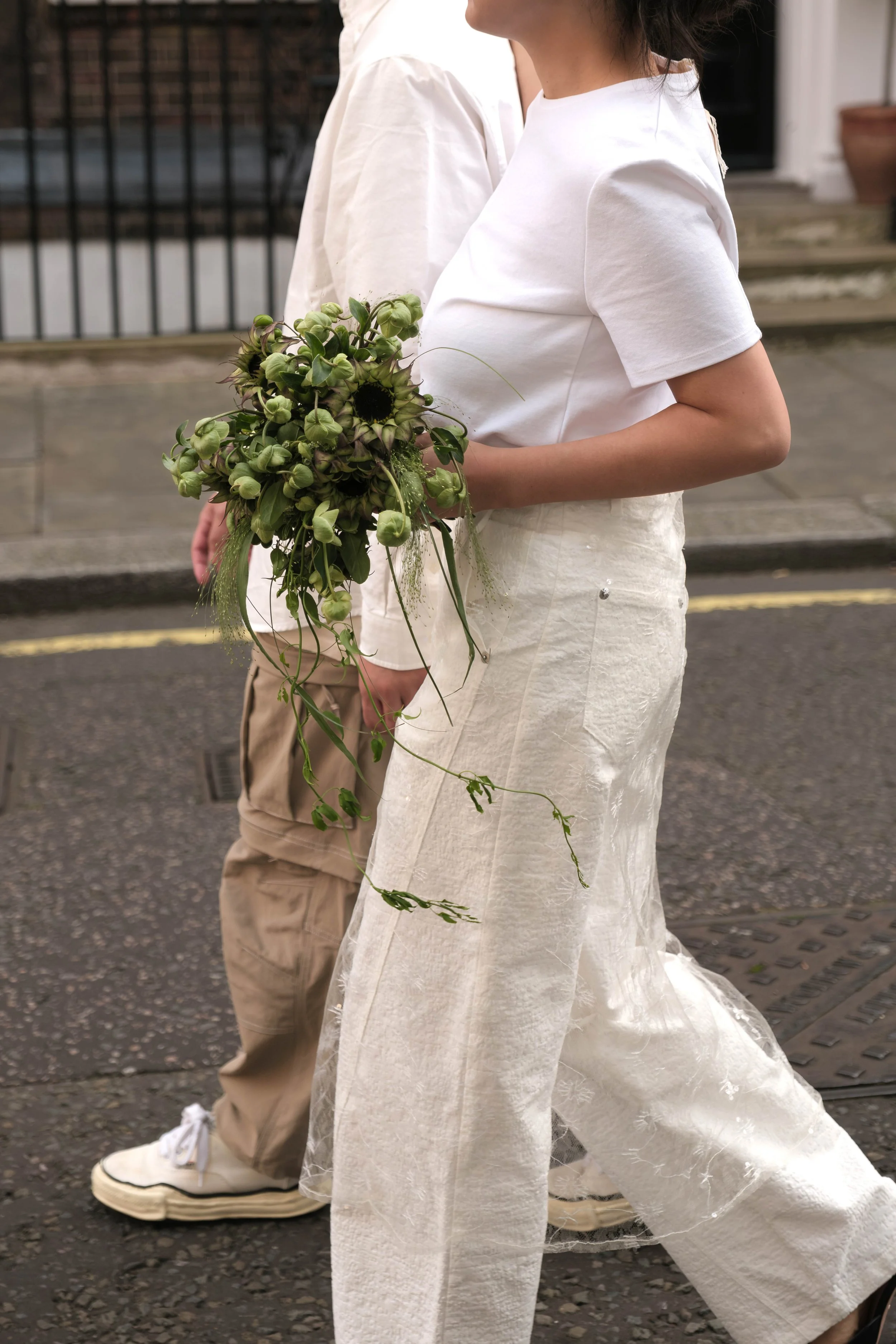 Person wearing white clothing holding a green floral bouquet walking outdoors on a sidewalk.