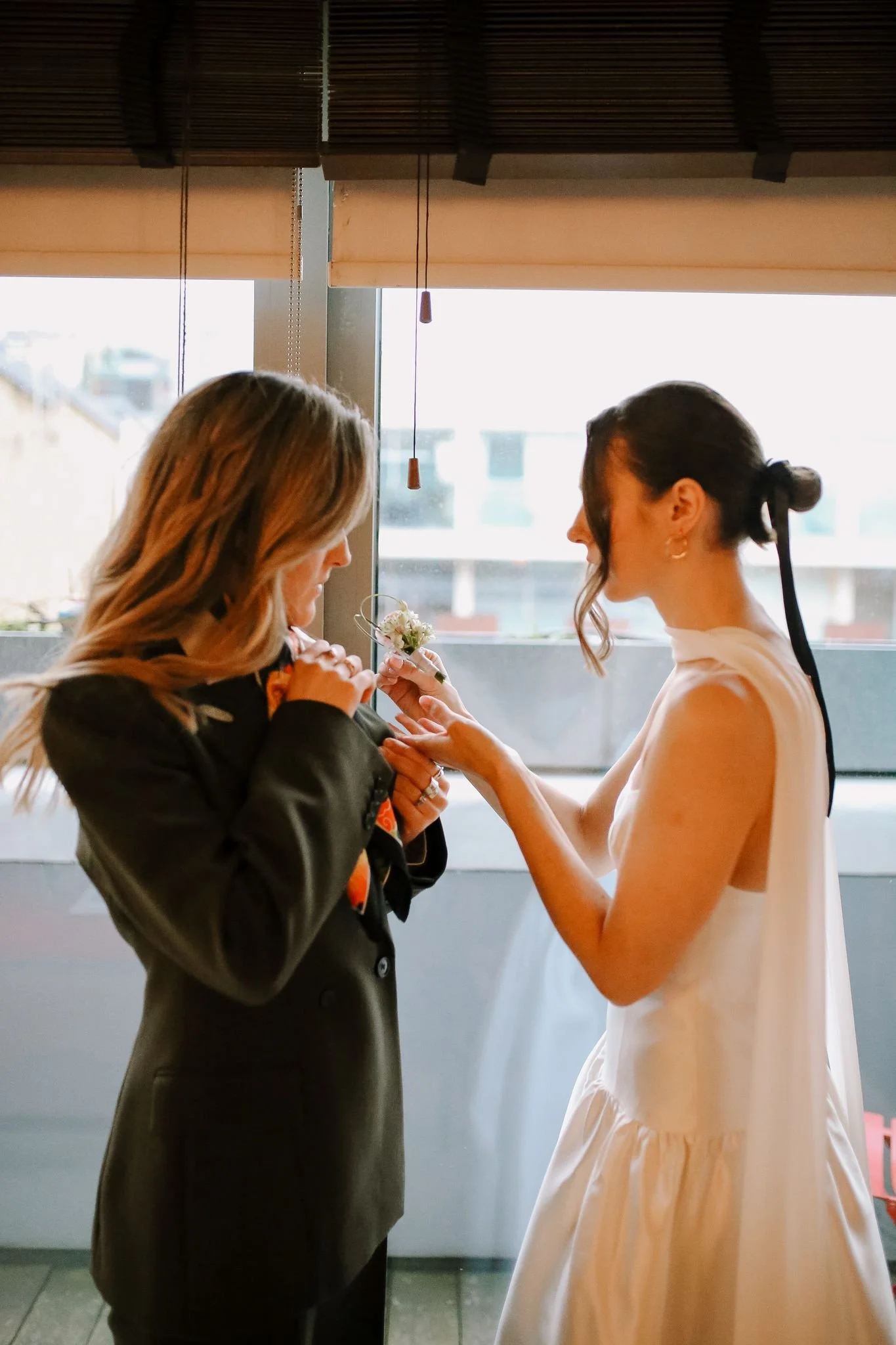 Two women are indoors near a window. One woman, dressed in a white dress, is helping the other woman, dressed in a dark suit, with a floral accessory on her coat.