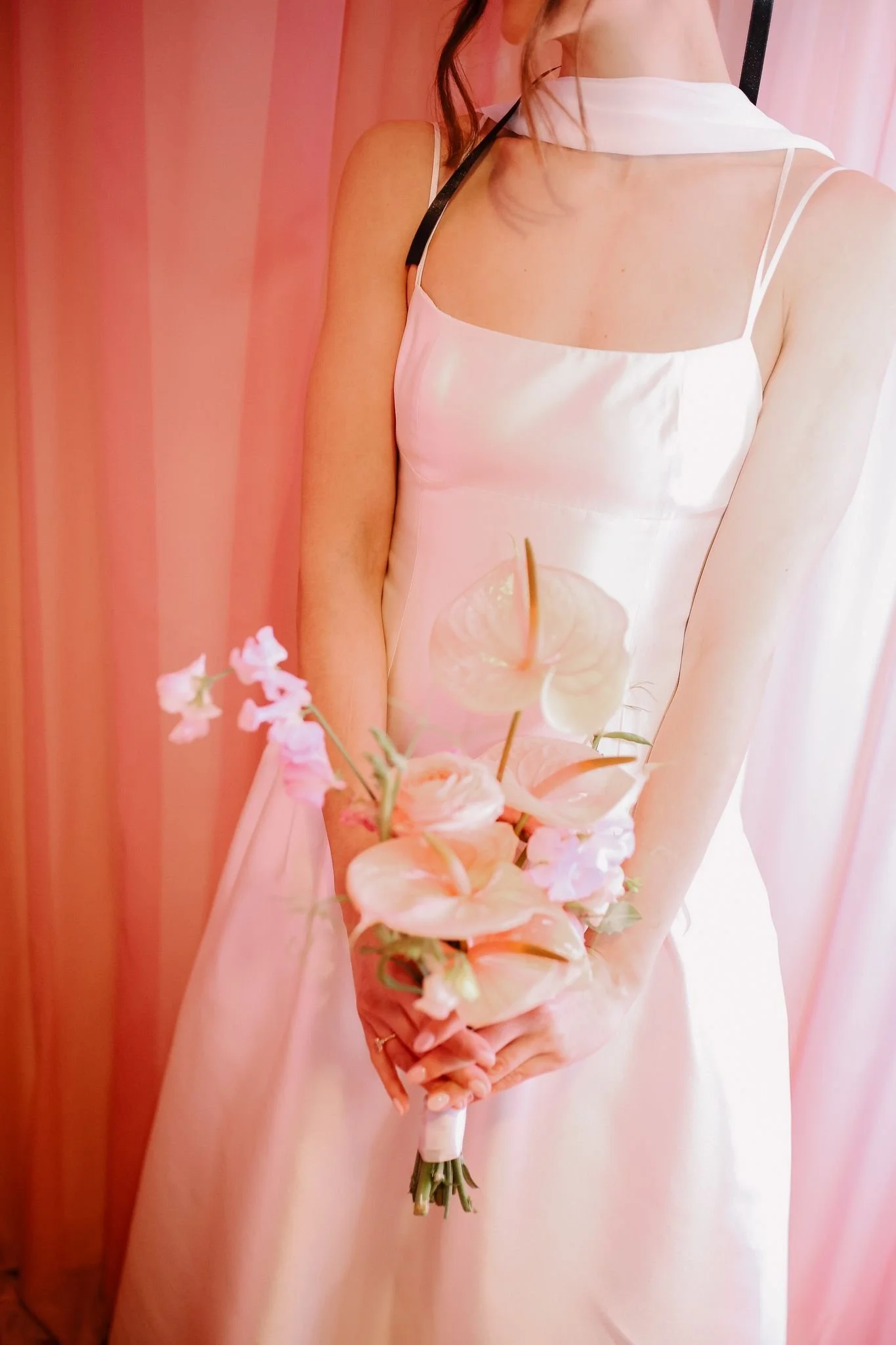 A woman in a silky, pale pink dress holding a bouquet of flowers. The background features pink curtains.
