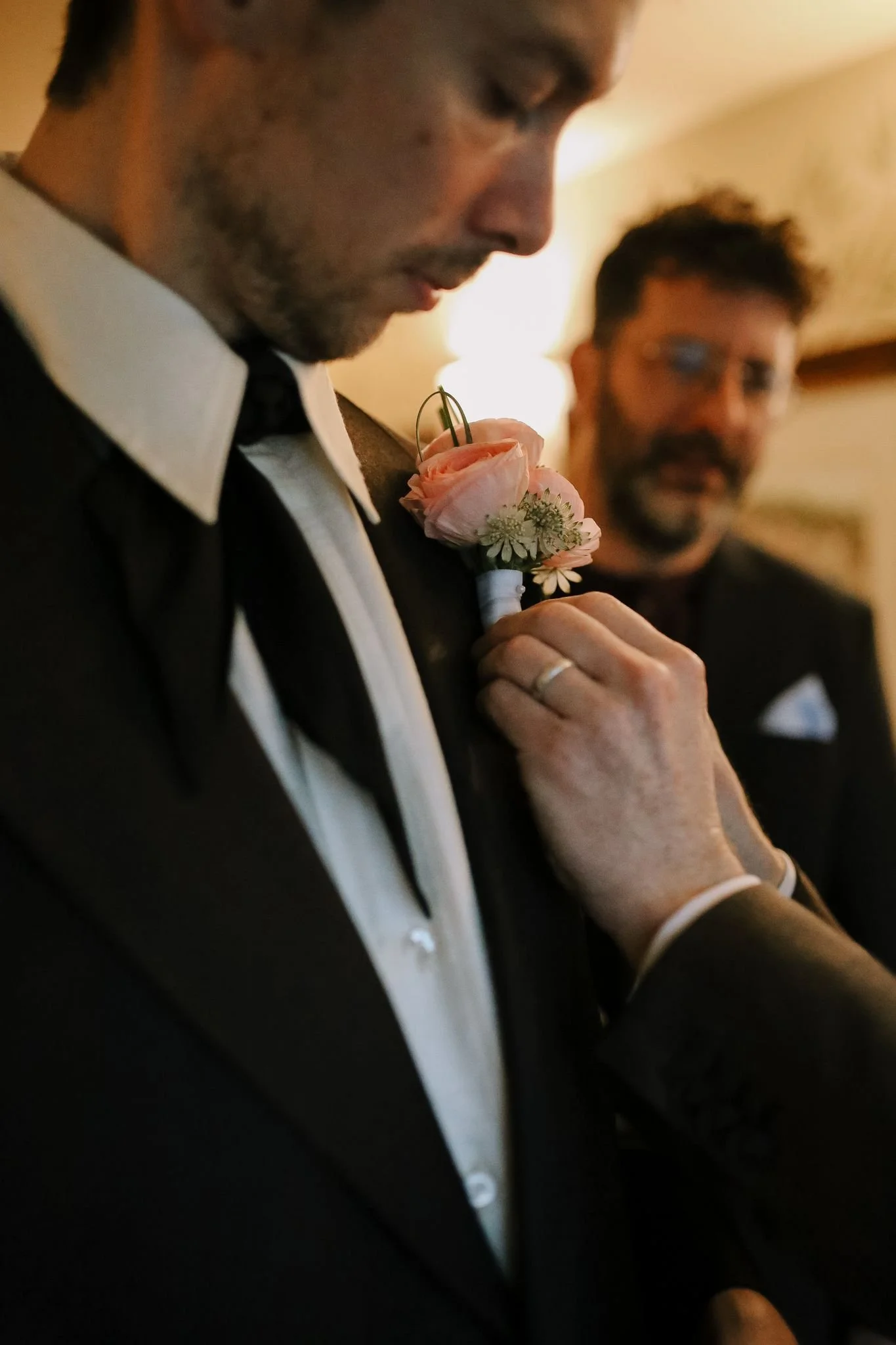 A man in a tuxedo with a boutonniere being pinned to his lapel by another person, with a second man in the background.