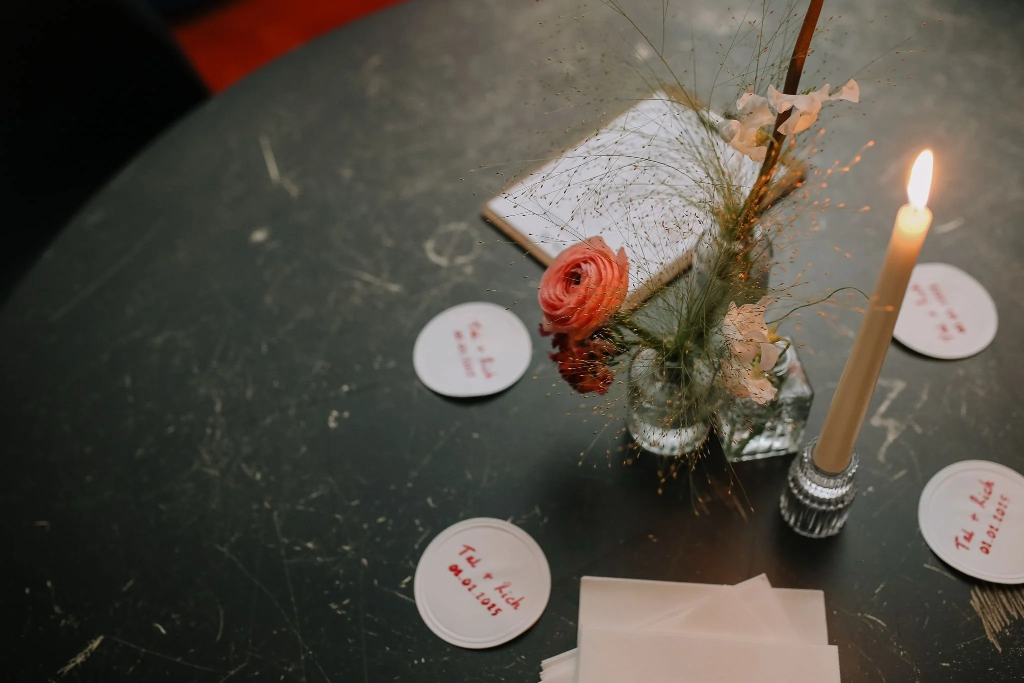 A dark round table with scattered white round paper coasters, a glass vase with dried flowers including a pink ranunculus and delicate grasses, and a lit beige taper candle in a decorative holder.
