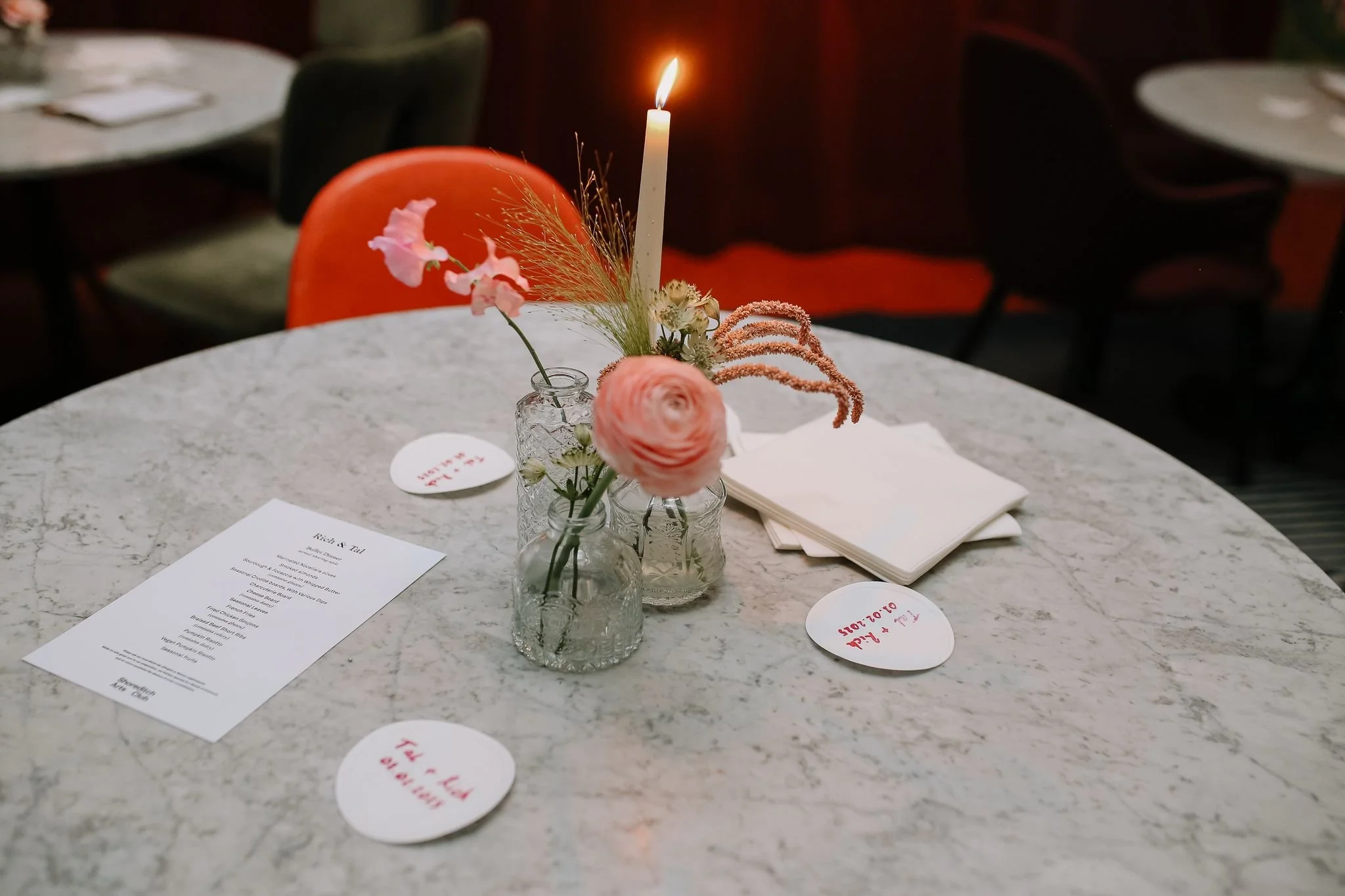 Table setting with small glass vases holding pink and white flowers, a tall lit candle, white napkins, and heart-shaped place cards with red and pink writing, on a marble table at a restaurant.
