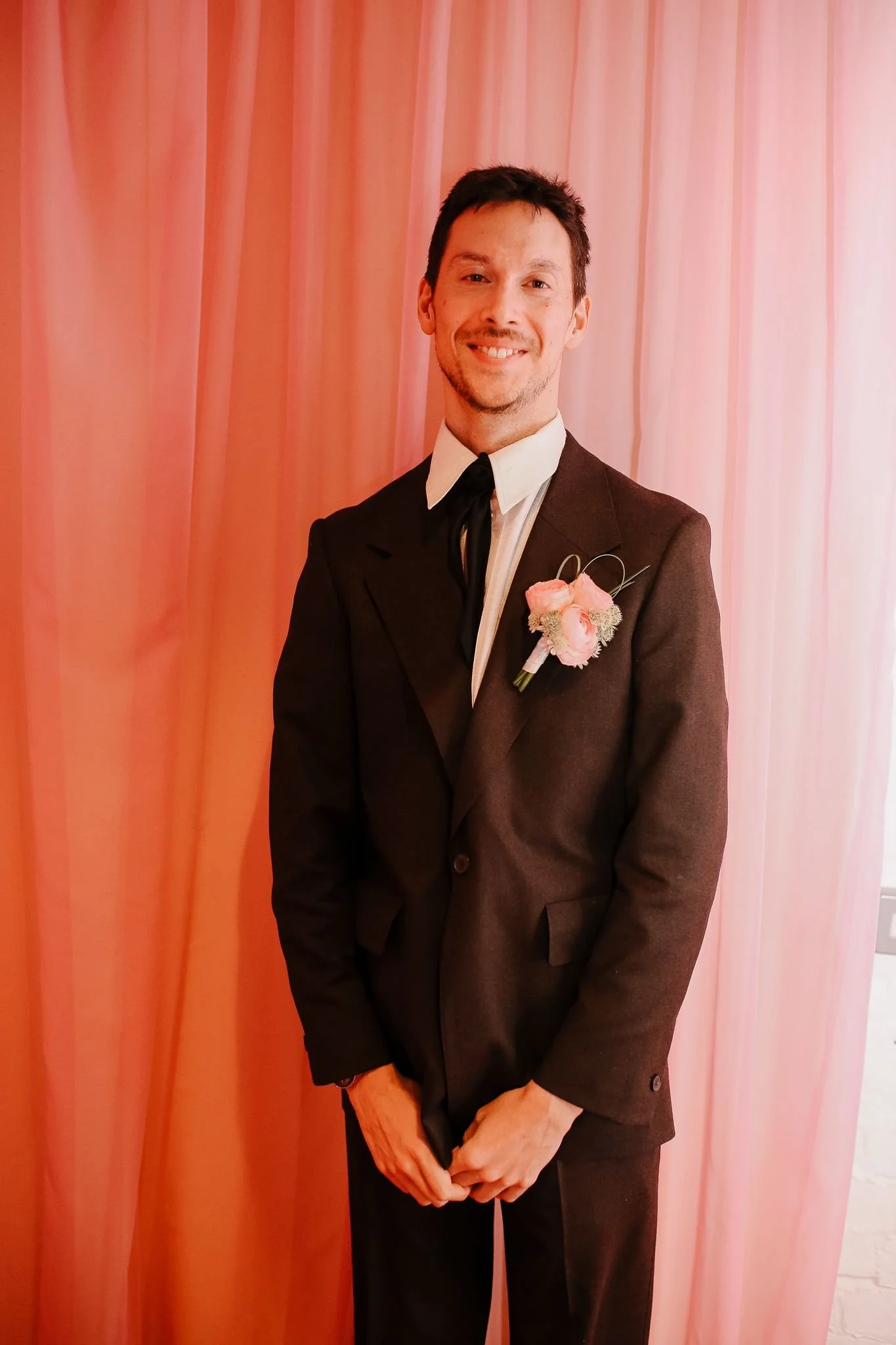 A man dressed in a black suit with a white shirt and black tie, standing in front of pink curtains, with a pink boutonnière on his left lapel, smiling at the camera.