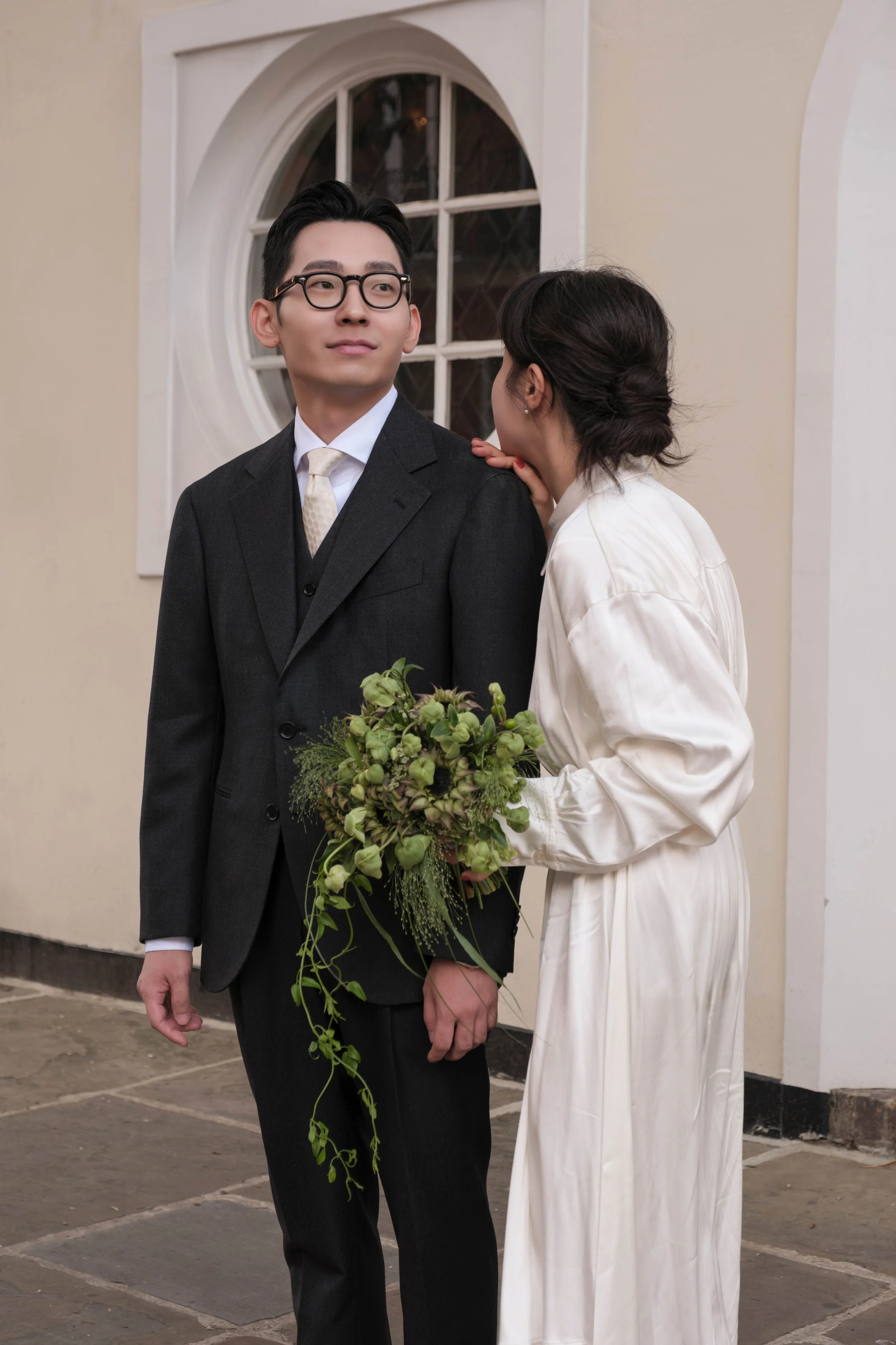 A woman in a white satin robe is whispering to a man in a dark suit while holding a bouquet of green flowers and foliage, standing outdoors near a beige building with a window.