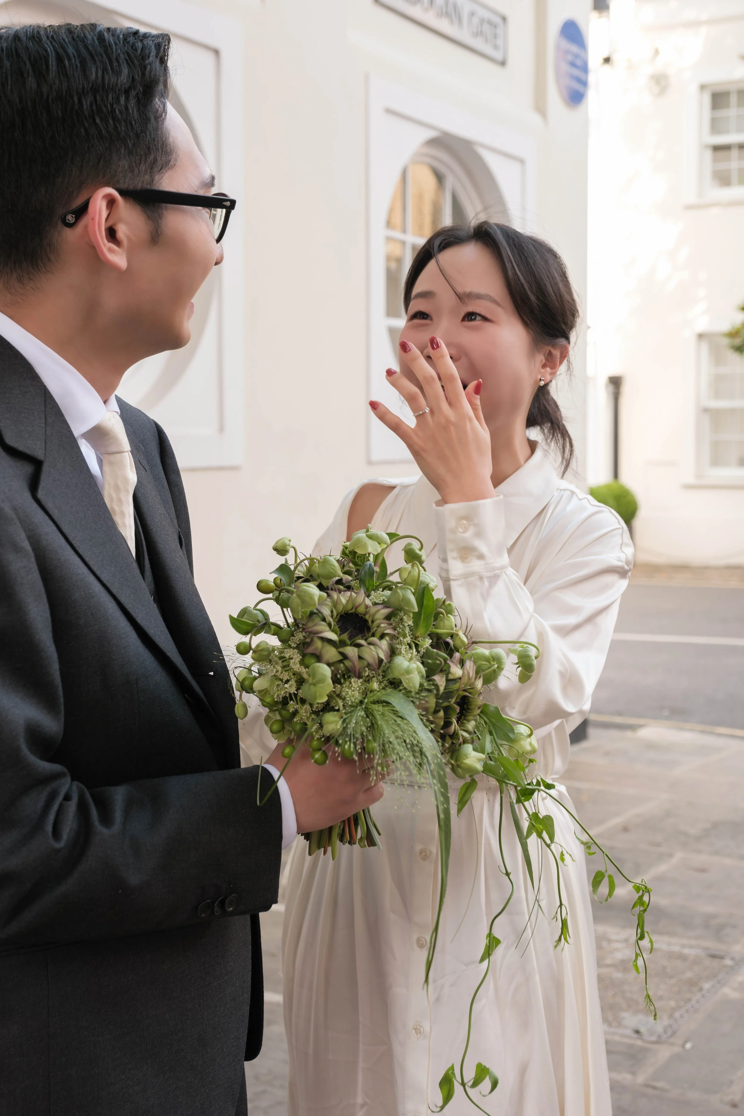 A man in a suit holding a bouquet of flowers is smiling and talking to a woman in a white dress, who is covering her mouth with her hand and appears emotional outside a white building.