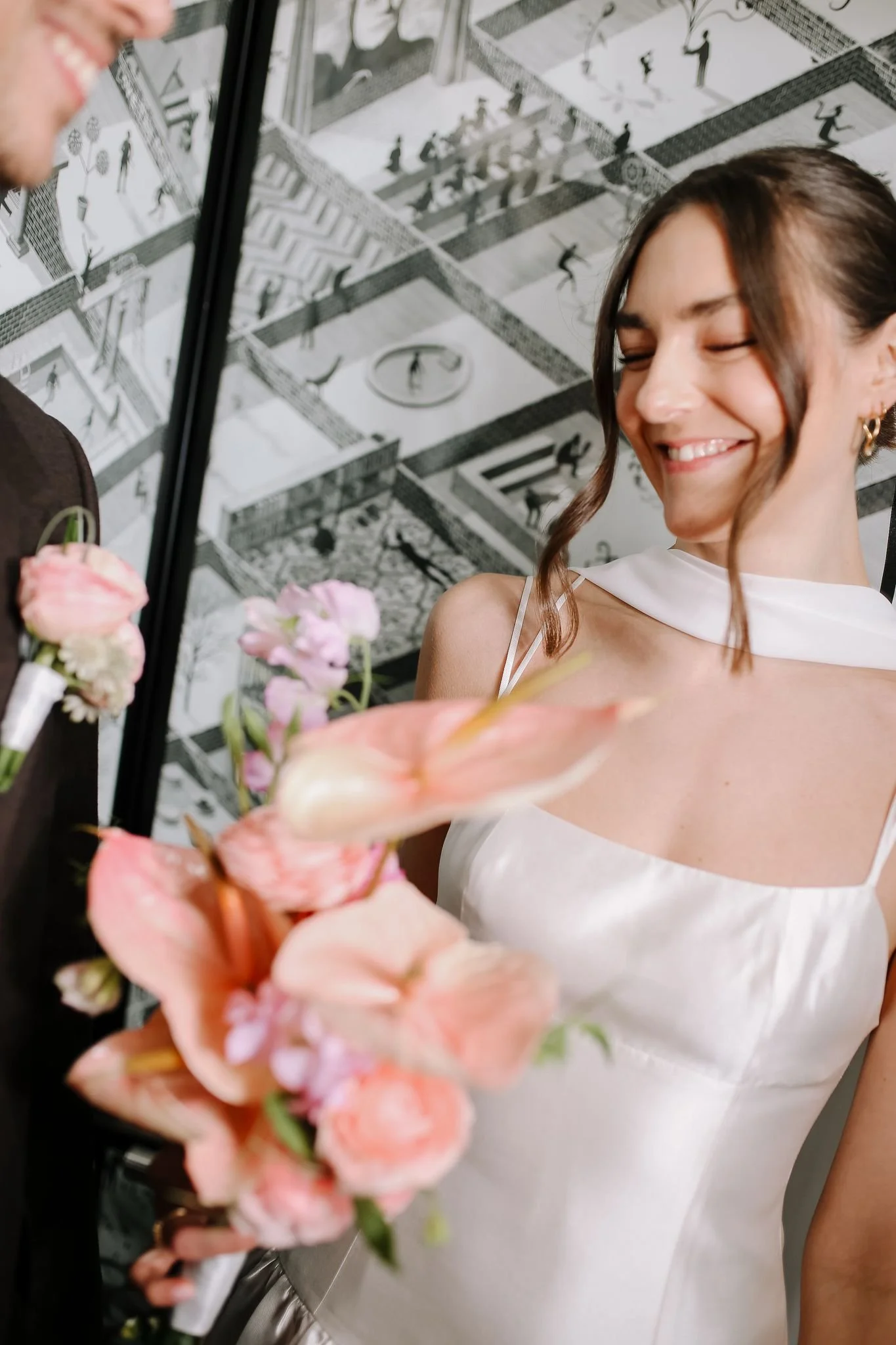 A bride smiling with eyes closed, wearing a white dress, holding a bouquet of pink flowers, standing near a man in a tuxedo with a boutonniere, in front of a black and white mural portraying abstract figures.