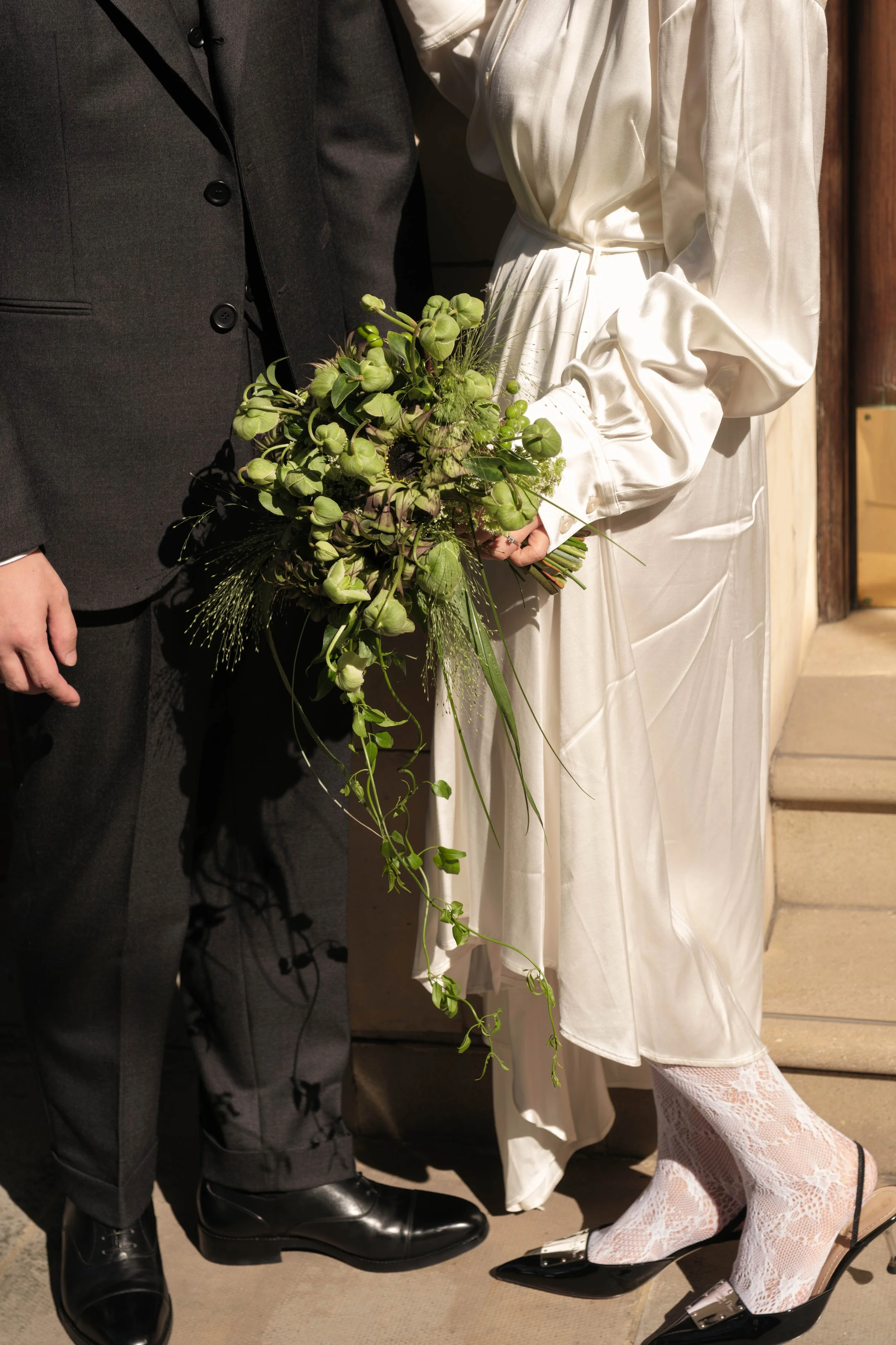 Close-up of a bride and groom holding a bouquet of greenery. The bride wears a white satin dress with lace stockings and black heels. The groom wears a dark suit with black shoes.