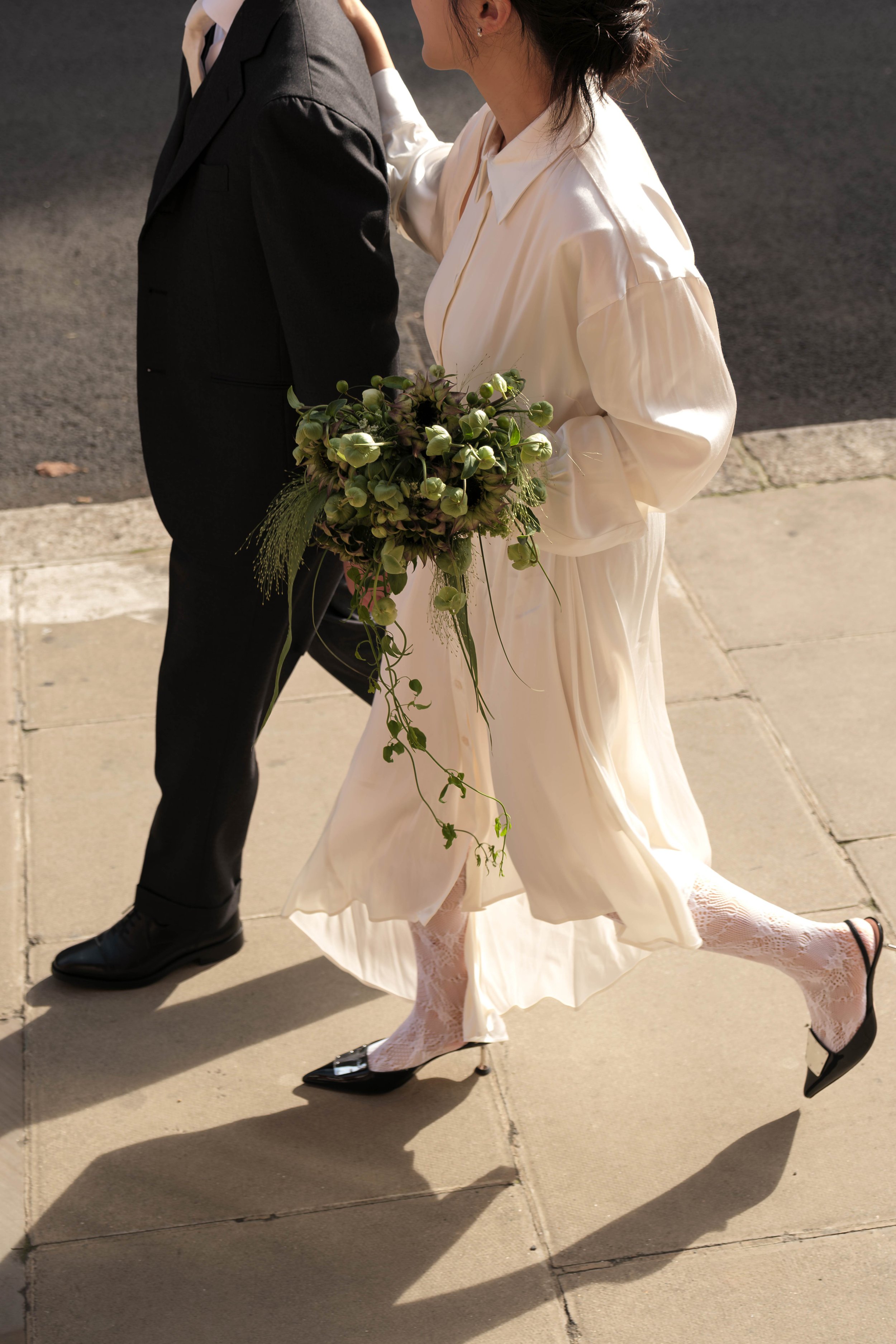 A woman in a cream-colored dress and lace tights holding a bouquet of greenery and flowers, walking beside a man in a black suit on a sidewalk.