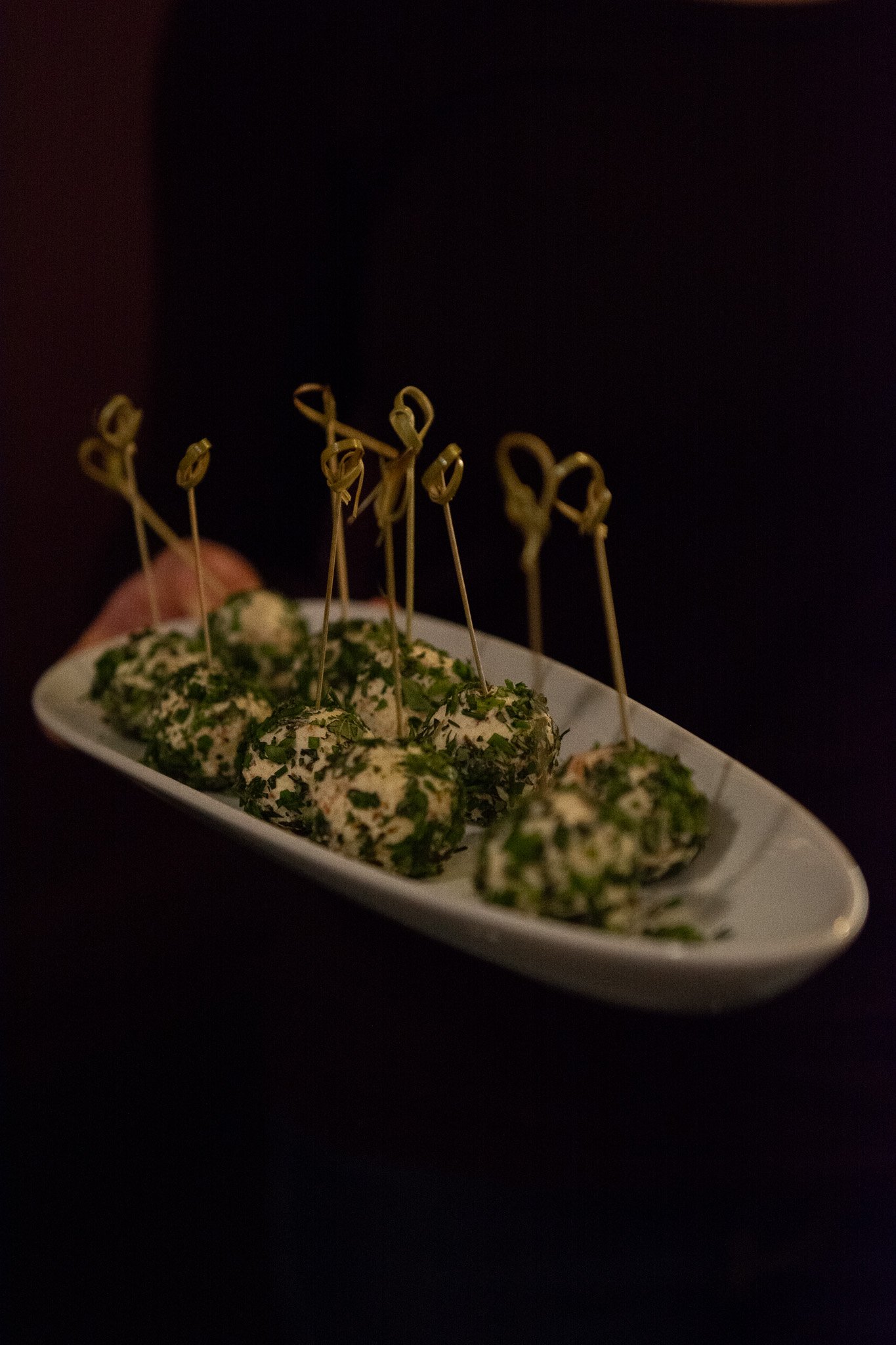 Jalapeno, bacon, cheese balls rolled in fresh herbs with tooth pick on white platter, hand holding platter