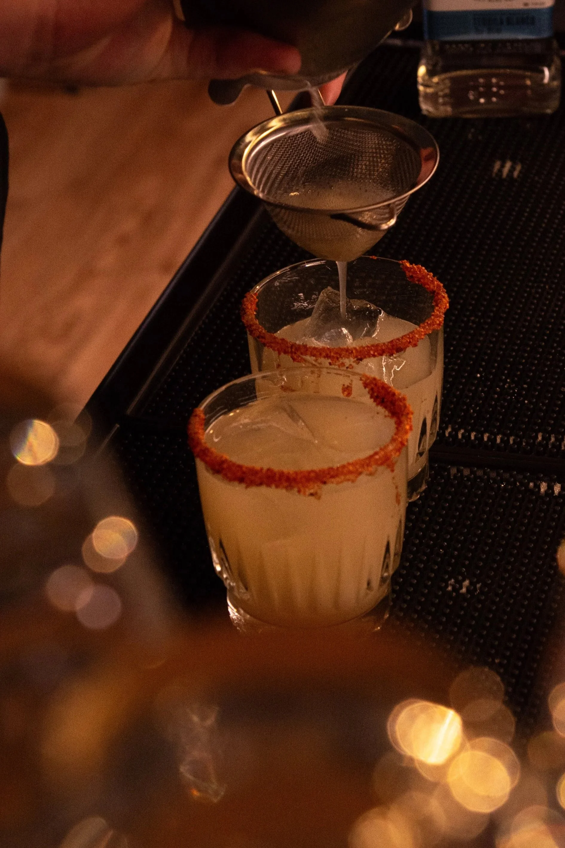 margarita with chilli salt rim being poured over ice through fine strainer with chopping board and hawthorne strainer in background